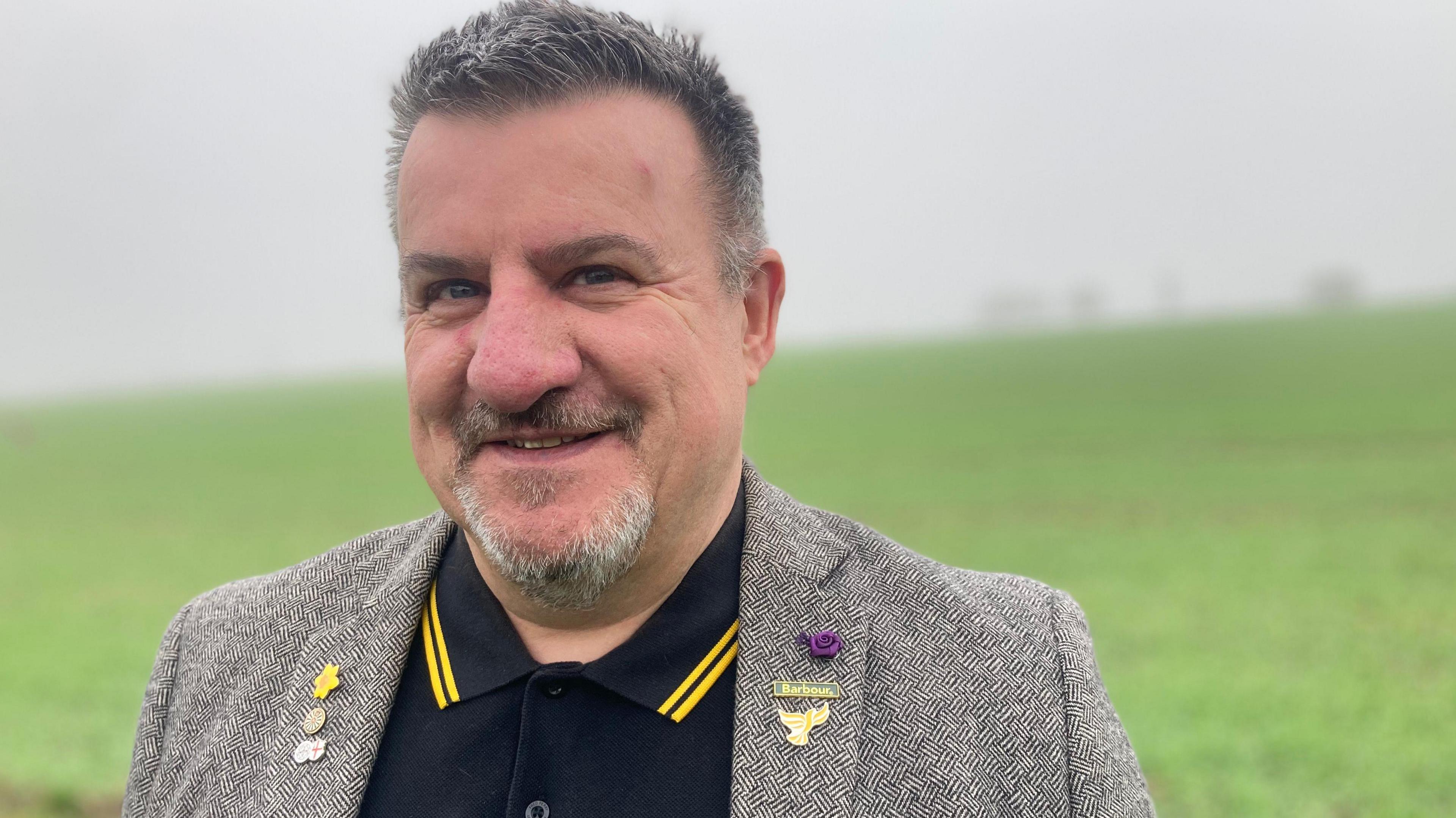 A head-and-shouolders shot of Des Fulcher, who has short grey hair and a grey, goatee beard and is wearing a grey jacket over a black top with yellow stripes on the collar. There are pins on the jacket.  