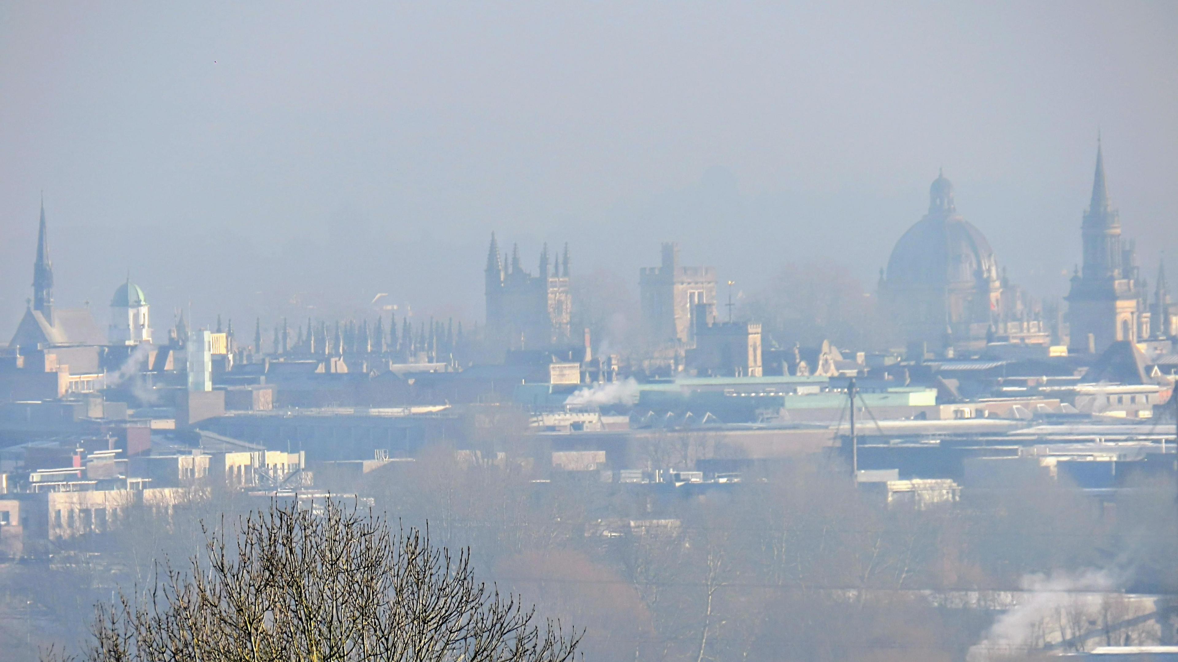 A city skyline can be seen on a hazy day. The spires of Oxford's old buildings are on the horizon against a misty sky with more modern buildings and a tree in the foreground.