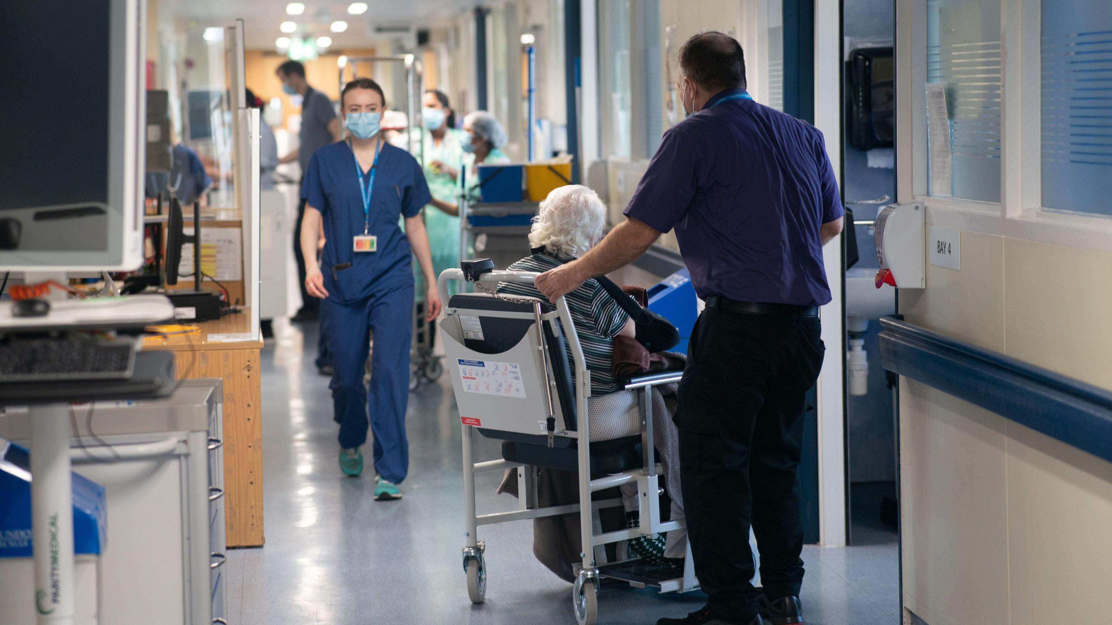 An NHS hospital worker, wearing a surgical mask and scrubs, walks down a busy hospital corridor 