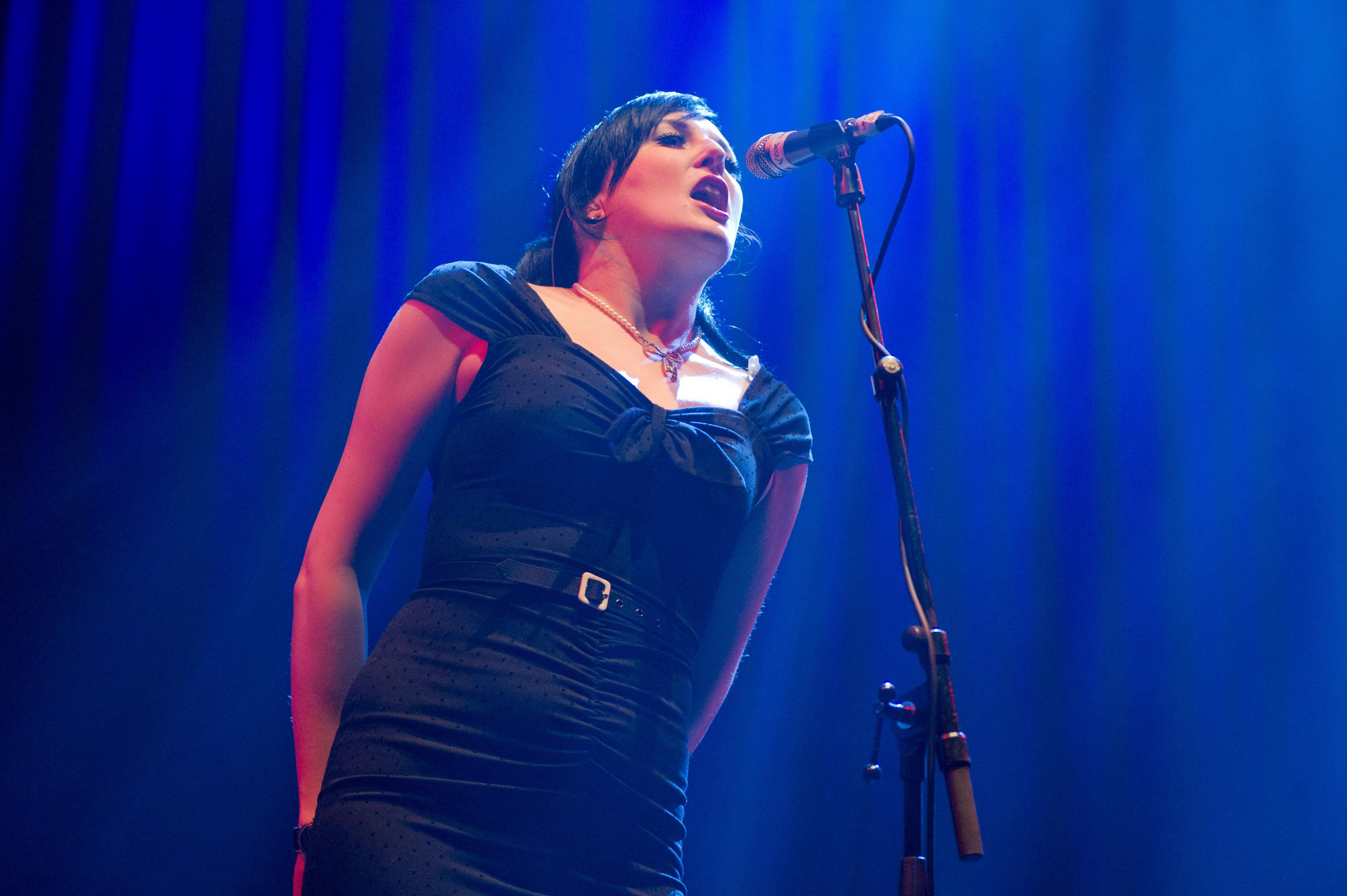 Lou Hickey sings onstage - she has dark hair and is wearing a black dress.