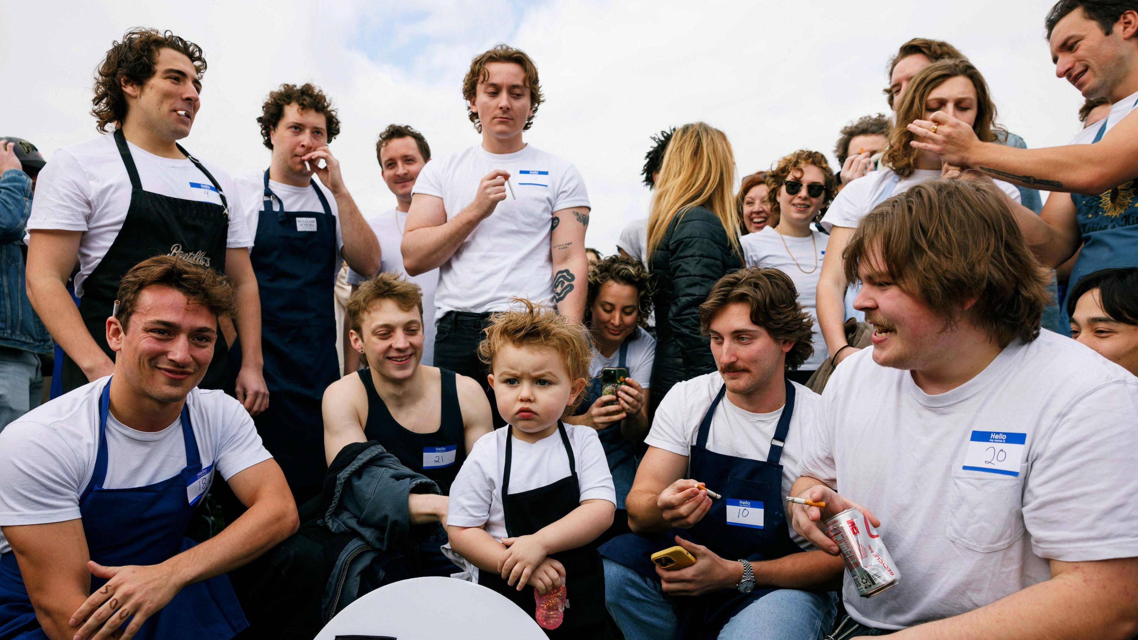 A crowd of Jeremy Allen White lookalikes, including a toddler, dressed in chef's aprons and white t-shirts, crouch and stand in a half-circle in a park in Chicago