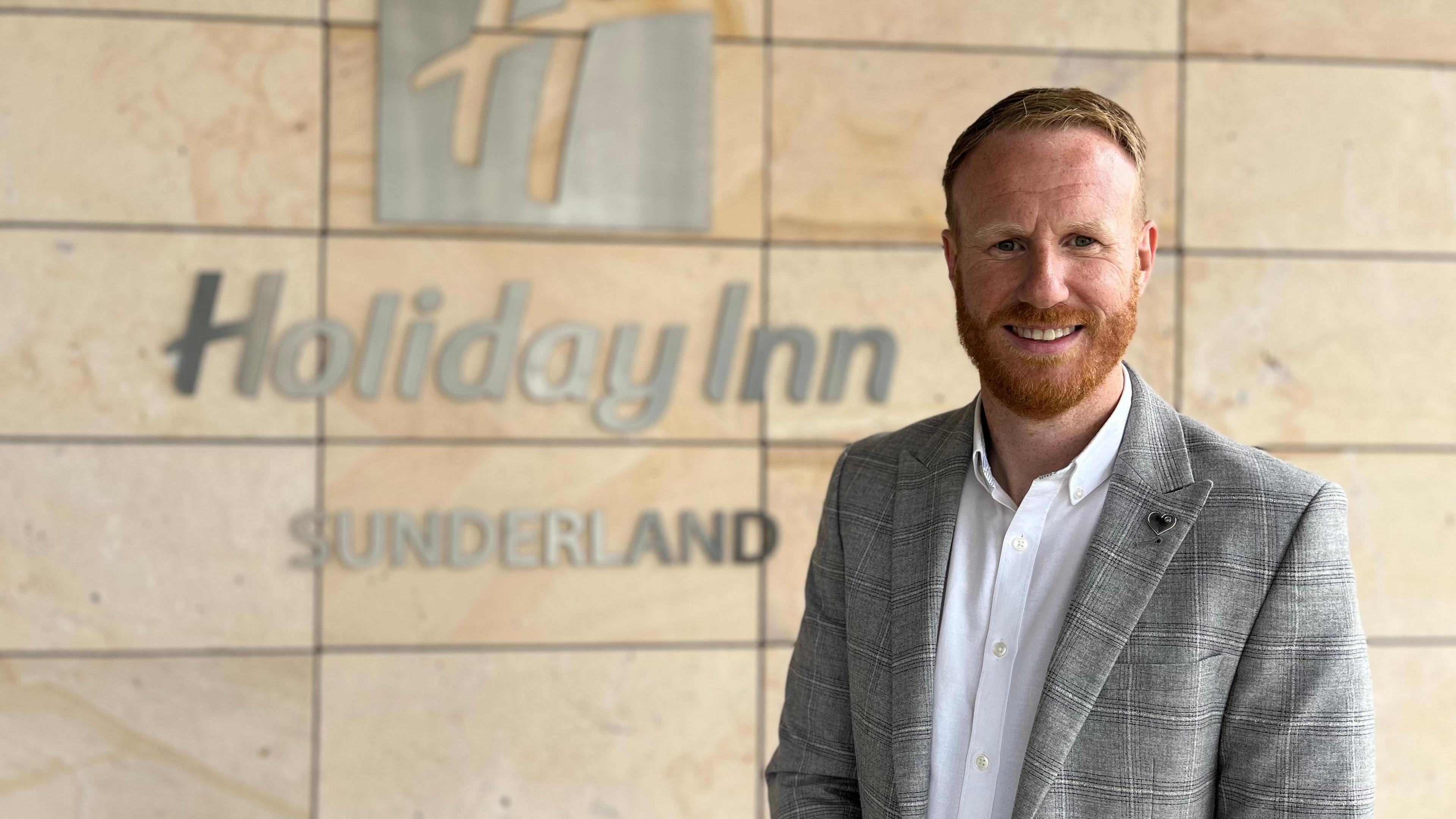 Lee Kirtland stands in front of a sign reading Holiday Inn Sunderland sign. He is dressed in a grey checked suit with a white shirt and has short red hair and a full beard. 