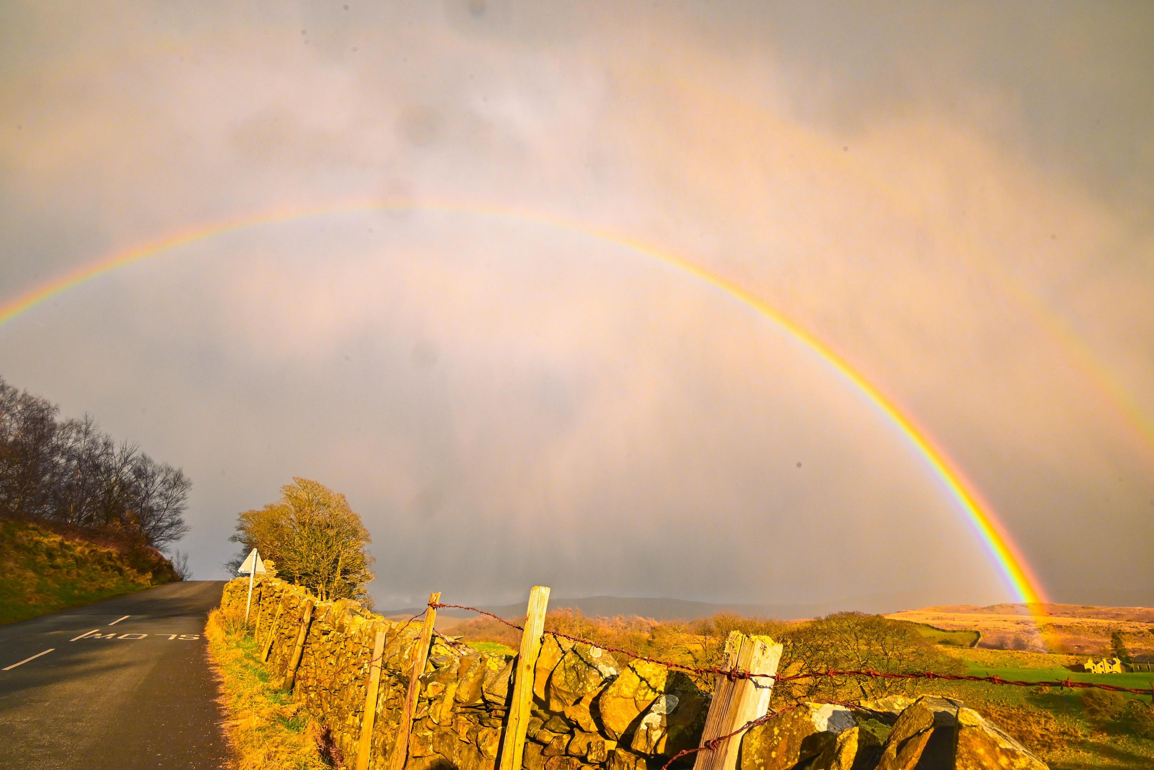 A rainbow over a rural road. A stone wall borders the road and a field.