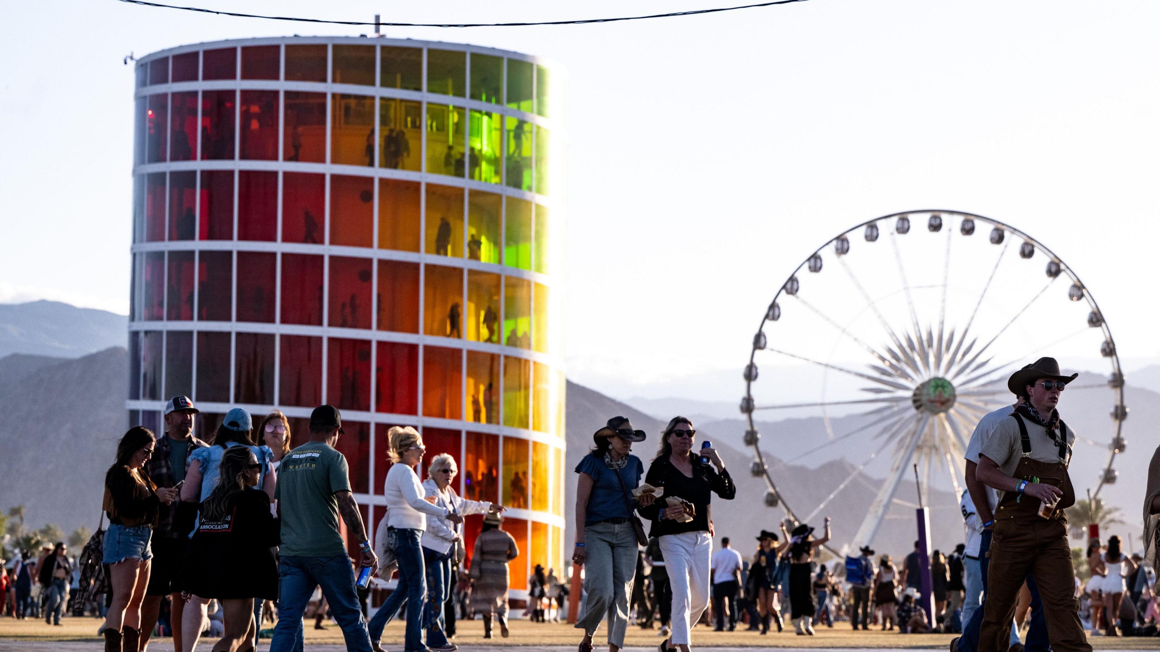 Crowds at the Coachella festival mingle in front of a large, cylindrical sculpture with red/green windows. A ferris wheel towers over the crowd in the background. Mountains fade away into the distance under a hazy sky.