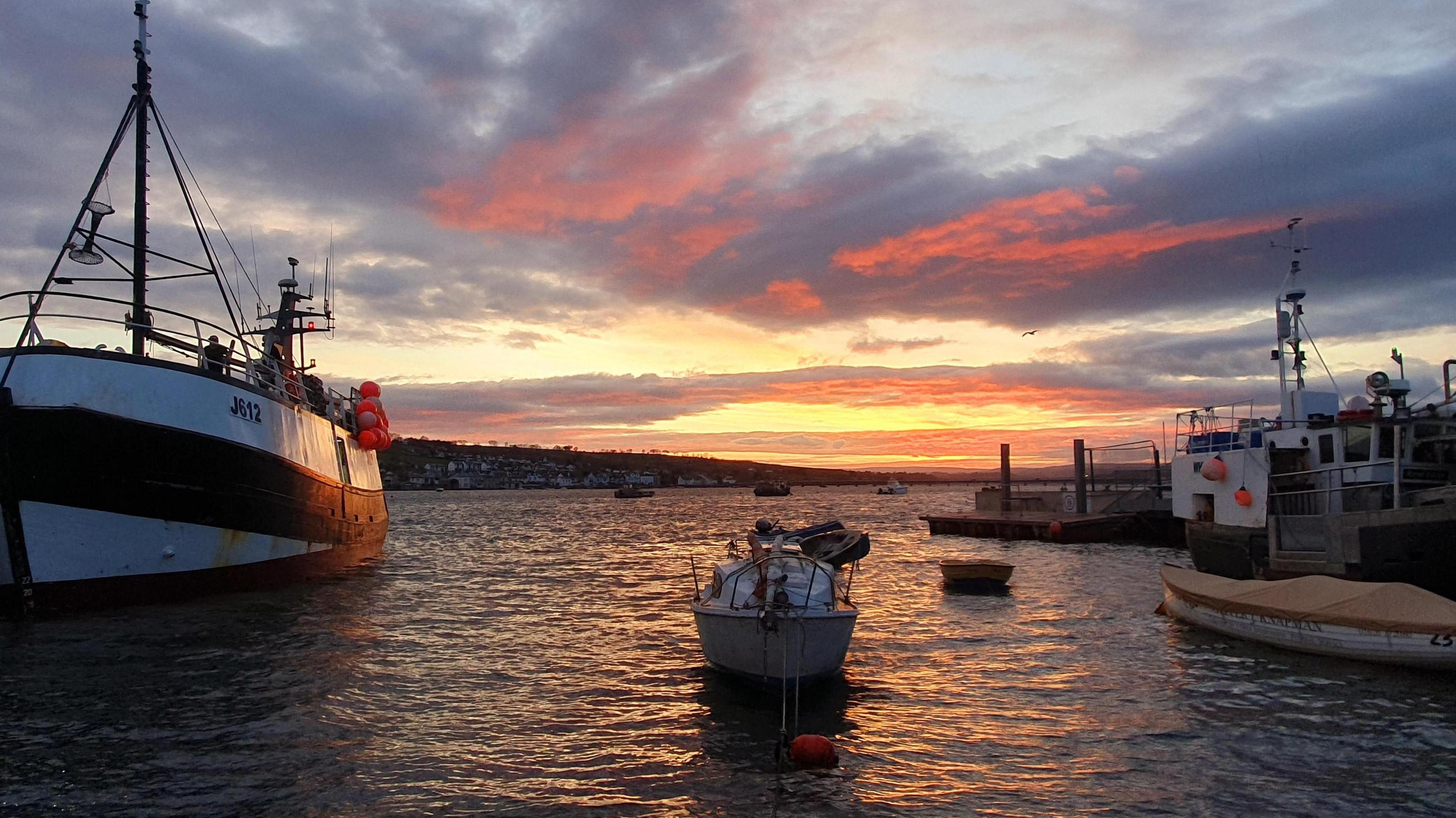 Three boats ranging in sizes bob on the water during a sunset in Teignmouth, Devon. A jetty is on the right-hand side of the shot.