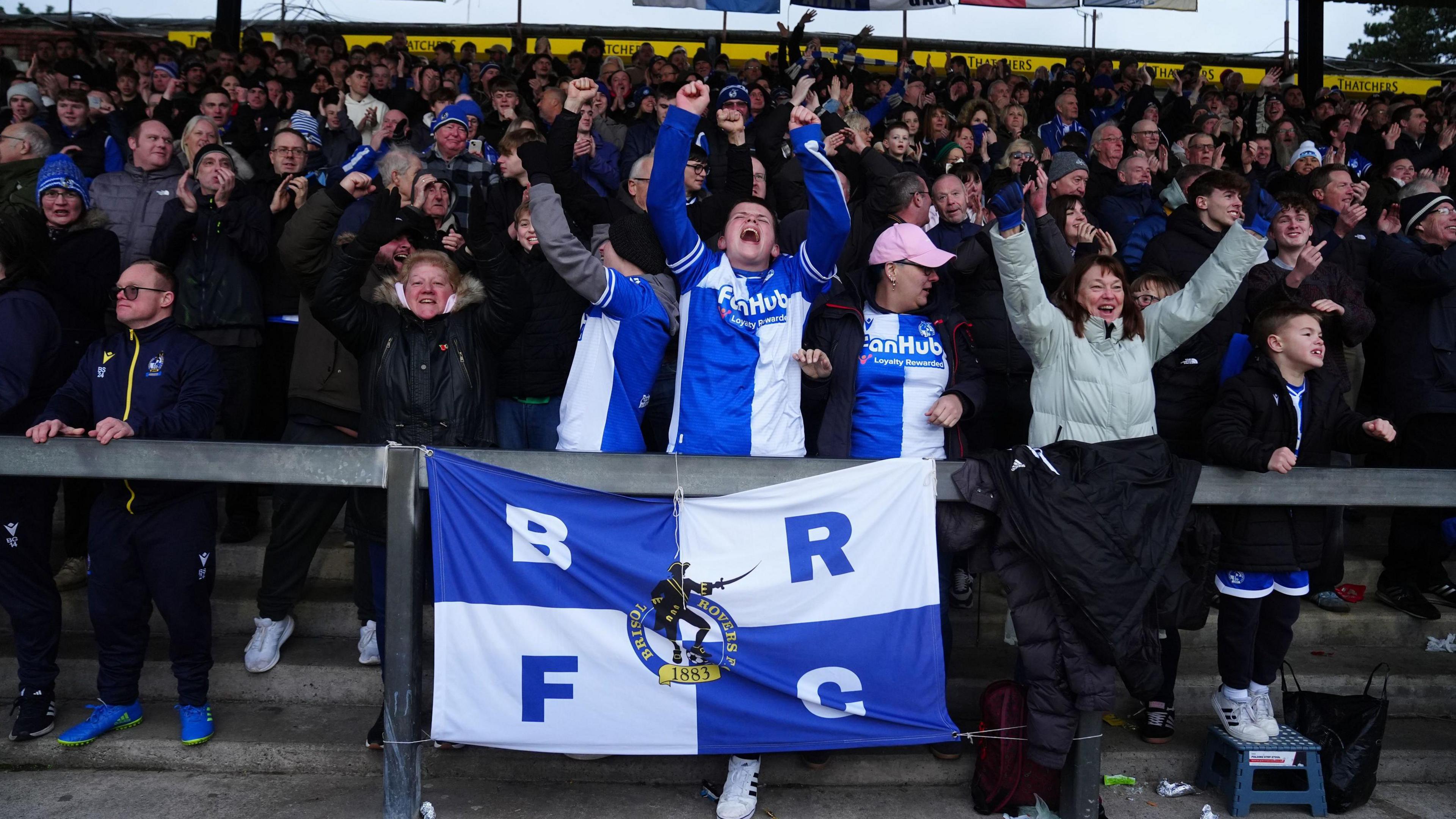 Bristol Rovers fans celebrate a goal in their game against Peterborough United at the Memorial Stadium. They are on a terrace and at the front is a large blue and white flag with "BRFC" in large letters and the club's crest on it