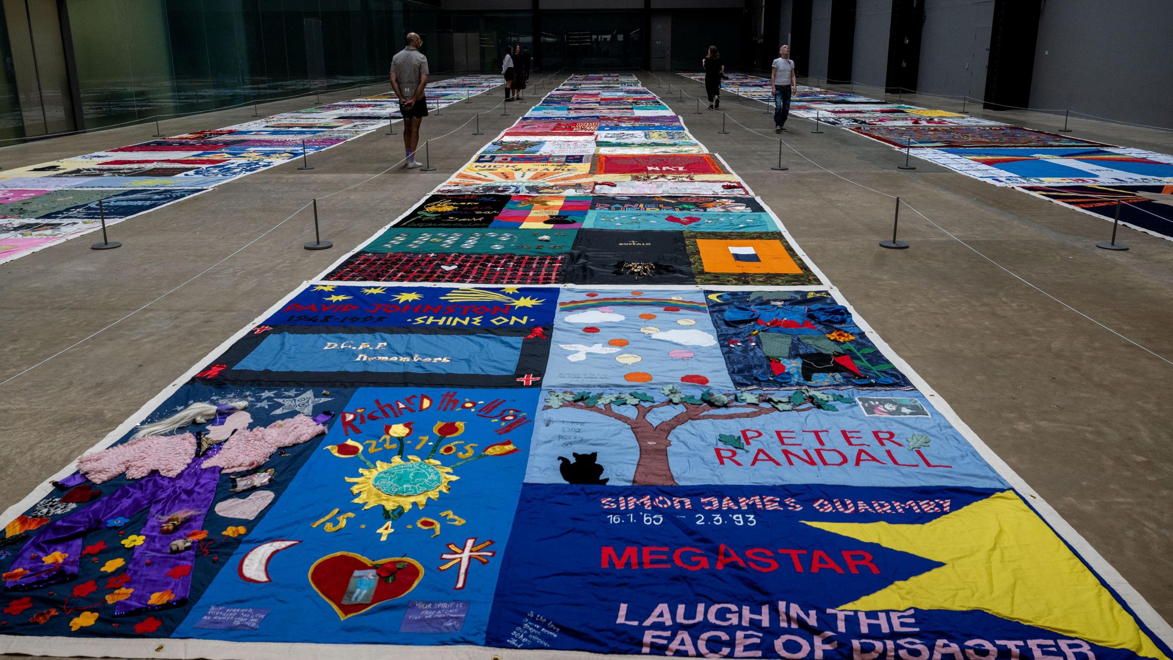 Closer image of a section of the UK AIDS quilt shows some of the individual submissions. One has a large tree and a cat, another the name of its subject and a quote, and another has some appropriate symbols.