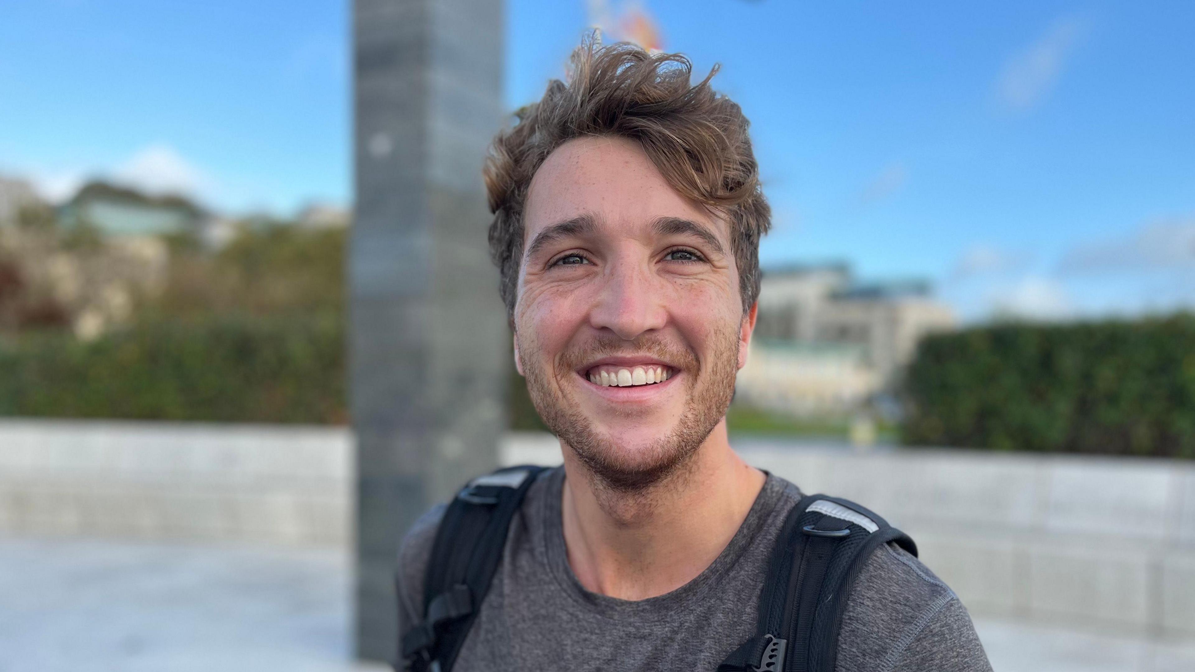 A man with medium length wavy hair with a backpack on smiling at the camera.