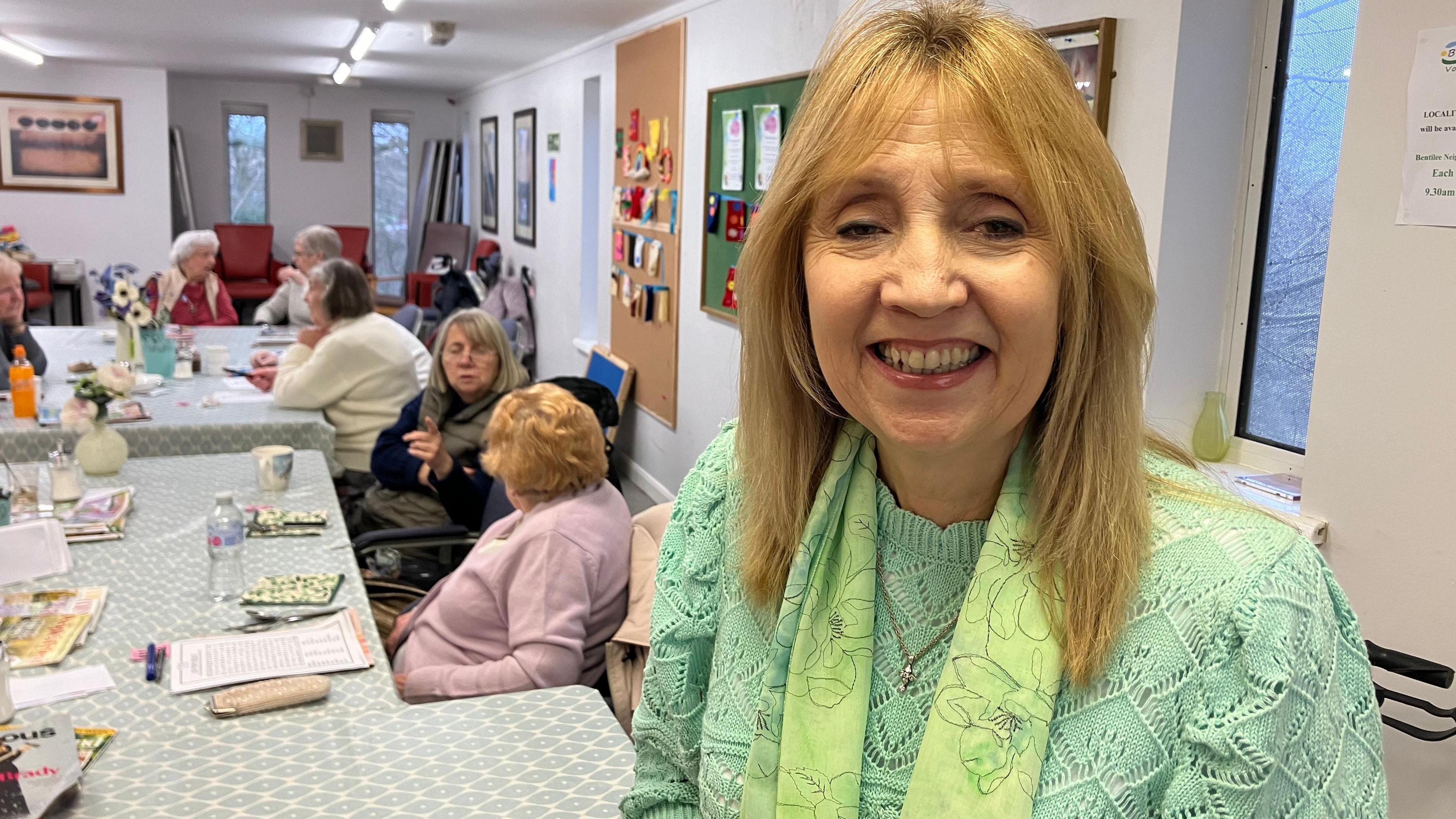 A woman with long blonde hair and wearing a green scarf and top smiles. Behind her are a number of people sitting around tables talking.