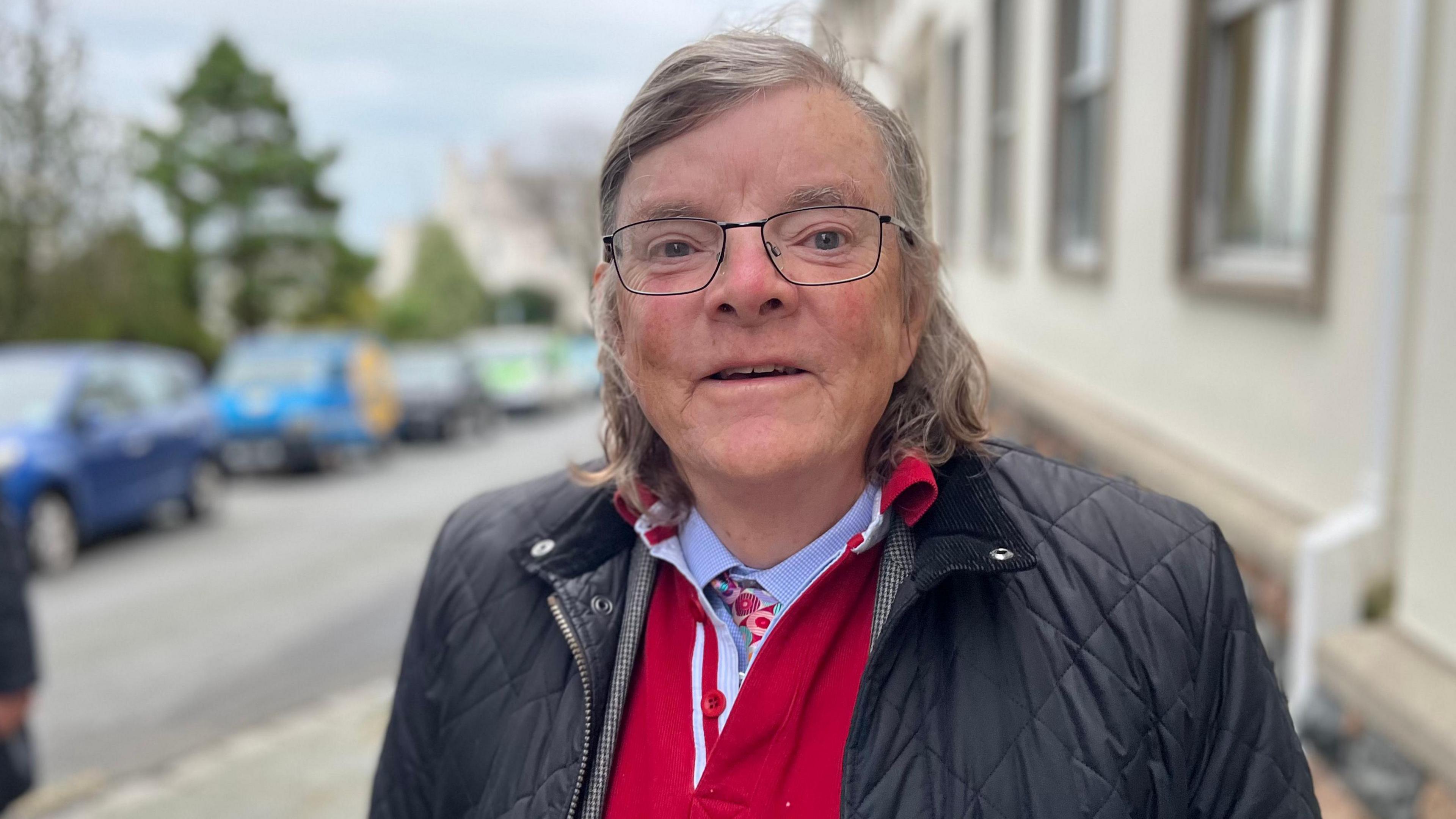 A man with grey and black shoulder length hair. He's wearing thin rimmed black glasses. He's wearing a blue Barbour jacket and a red jumper underneath.