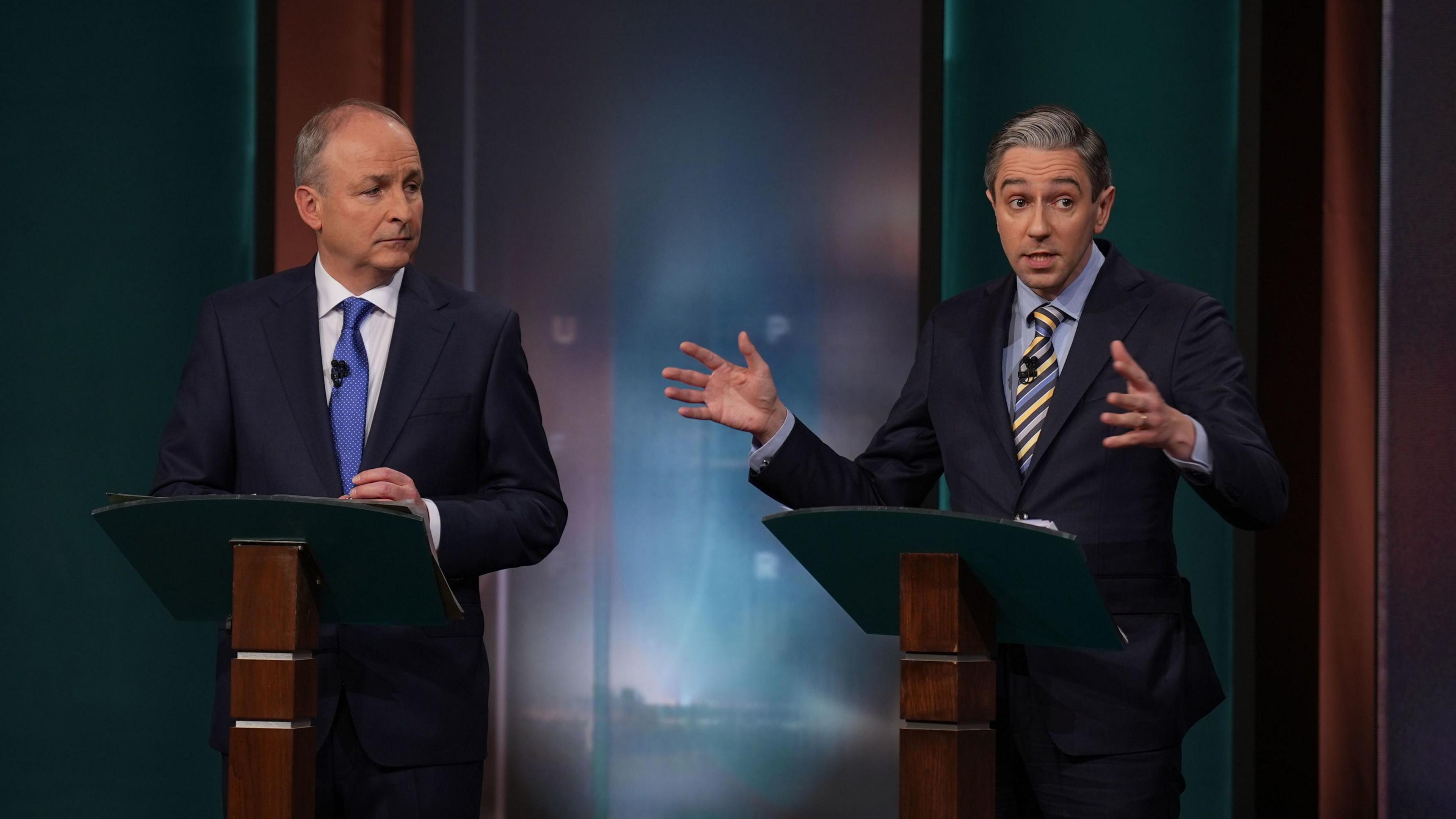 Micheál Martin and Simon Harris. Both are standing behind podiums made up of dark-wood and green. Both men are wearing dark suits. Martin is wearing a white shirt and blue tie while Harris is wearing a blue shirt and a blue and yellow striped tie.