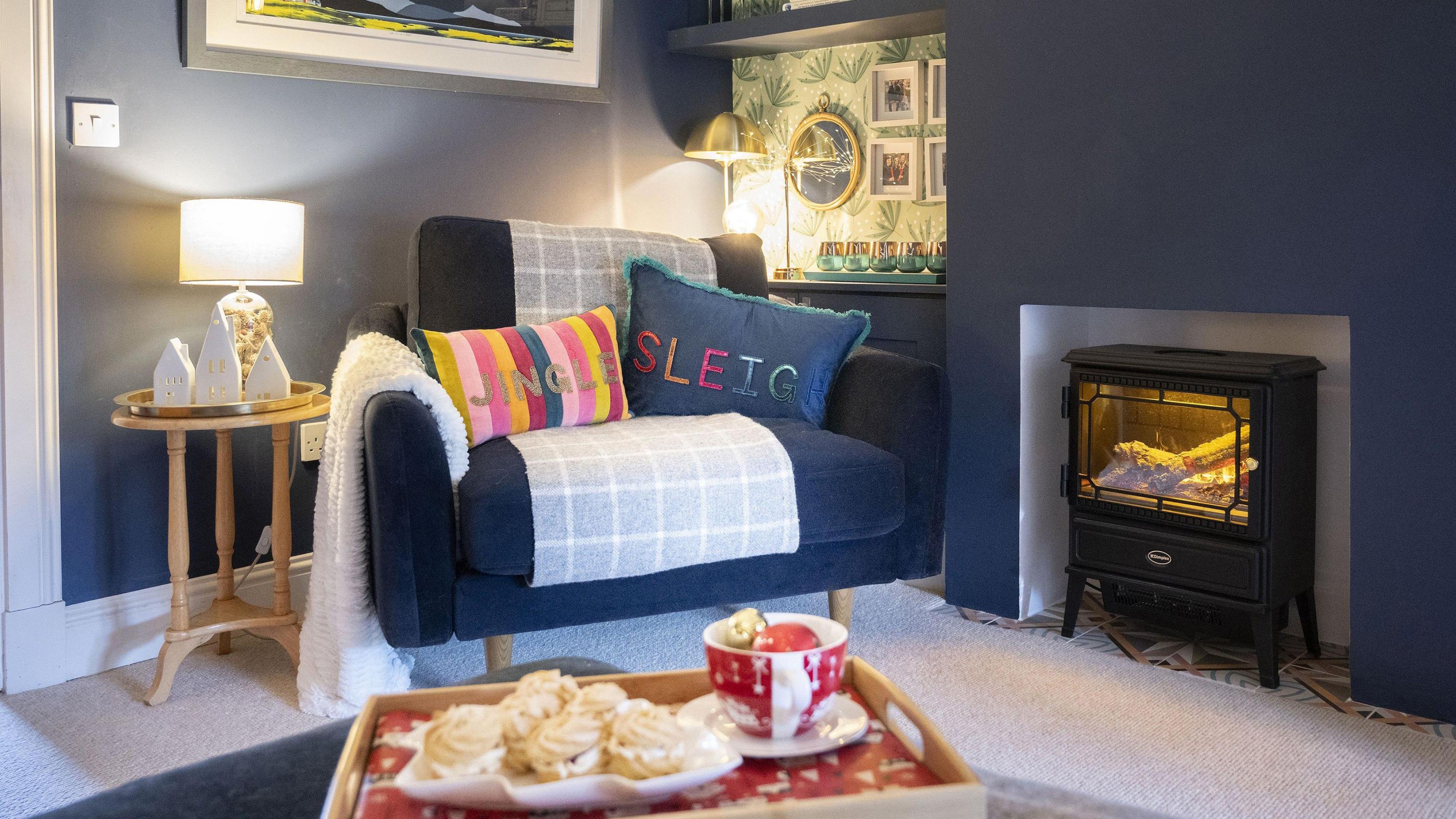 An armchair, decorated with two colourful pillows - one says 'jingle' and one says 'sleigh'. The armchair is situated next to a cosy fireplace. In front of the chair is a tray with a plateful of Viennese whirl biscuits.  