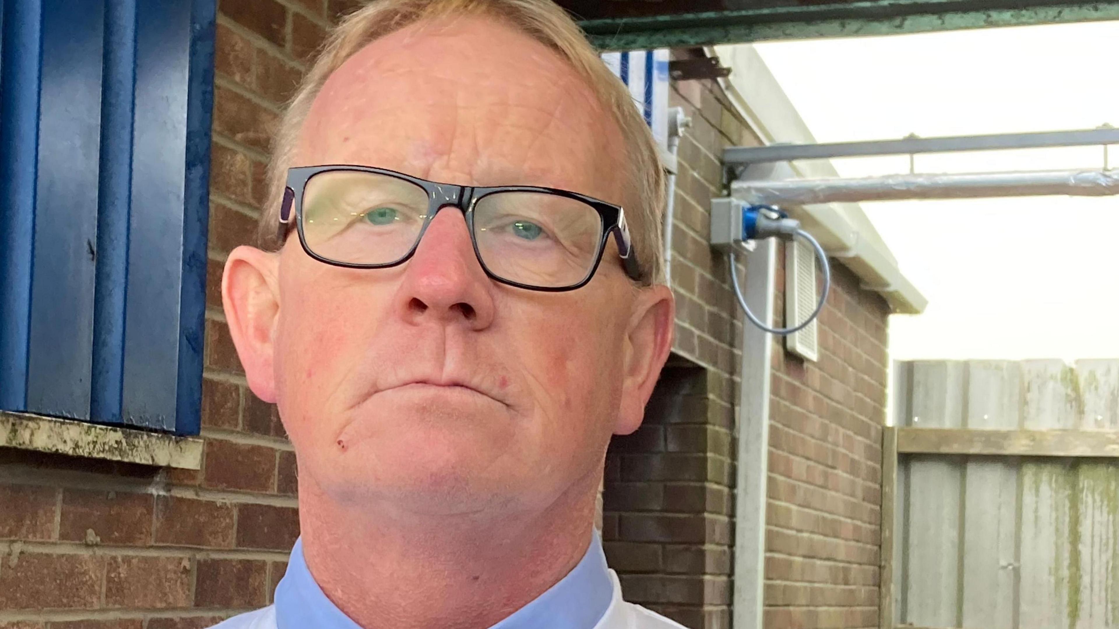 Jon Tindall, vice chairman of Bridlington Town FC, is wearing a formal blue shirt and dark rimmed glasses. He has short blond, greying hair. He is looking ashen faced.