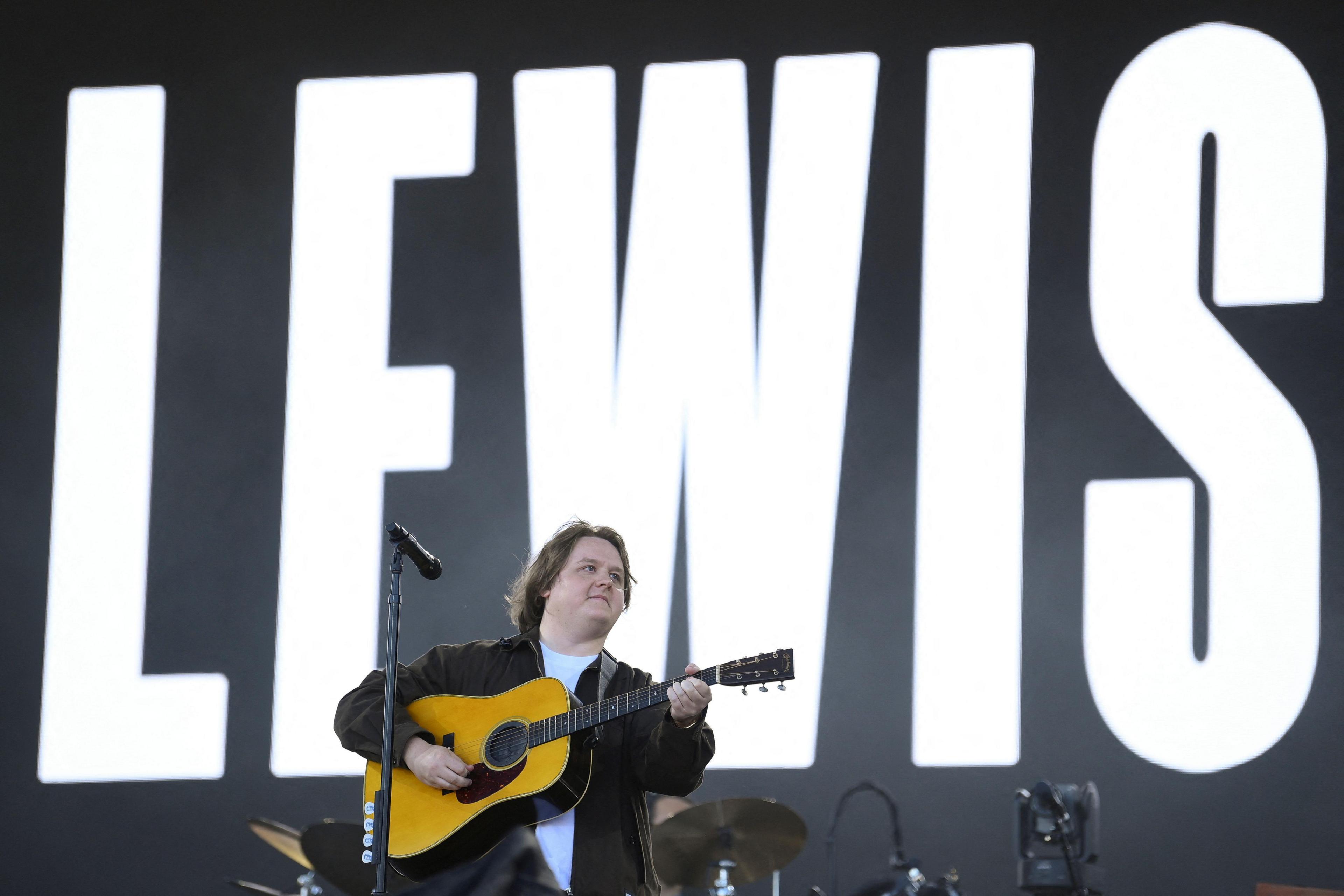 Lewis Capaldi strums an acoustic guitar on the Pyramid Stage at Glastonbury. He has lomng brown hair, with a middle parting, and is wearing a black jacket and white t-shirt. His name is projected on a screen behind him in the form of white lettering against a black backdrop.