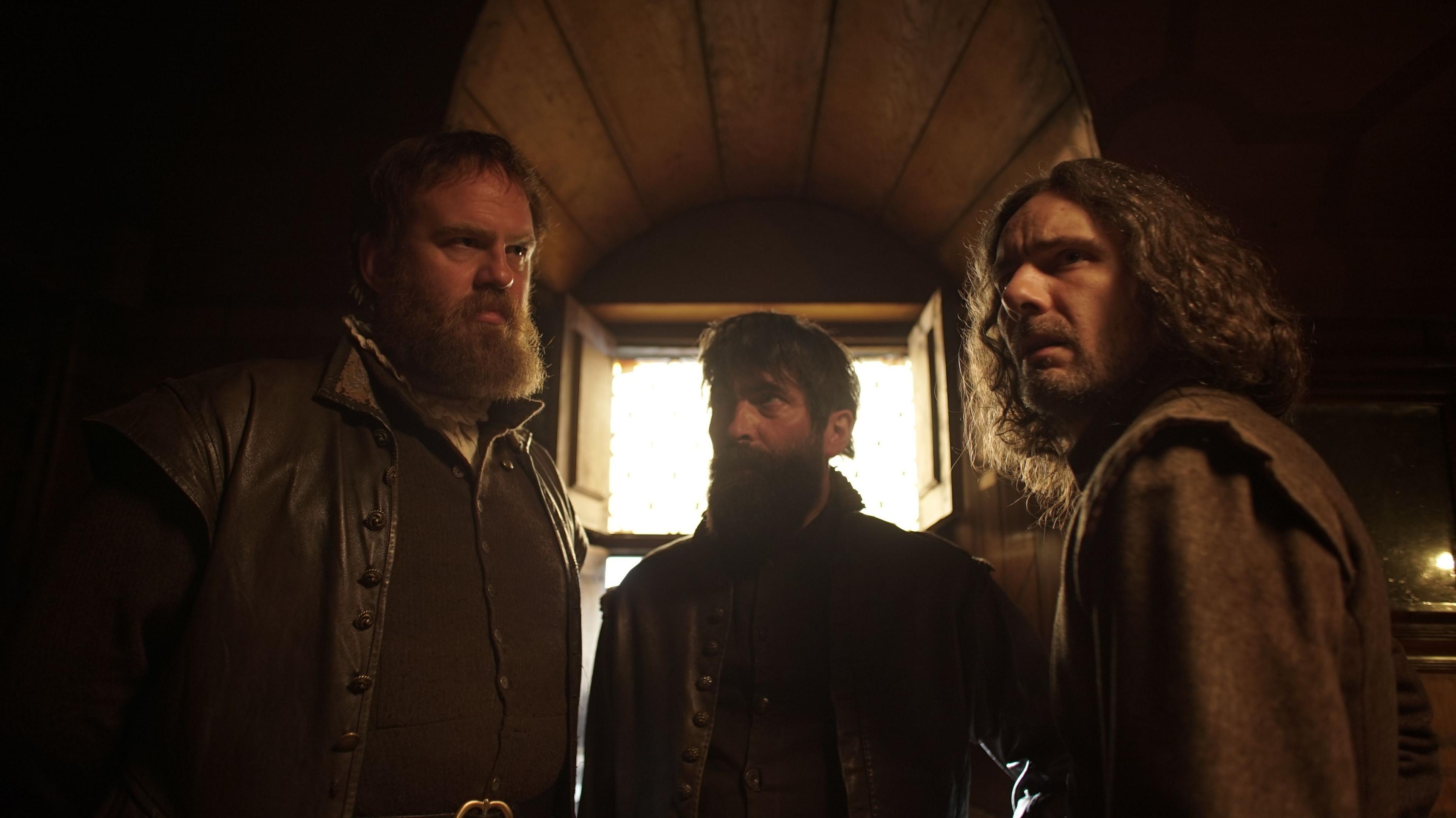 Three actors portraying Guy Fawkes, Thomas Wintour, and Robert Catesby. They are dressed in historical dark clothing, in a room, with a window, which is dark. They all look tense and serious. 