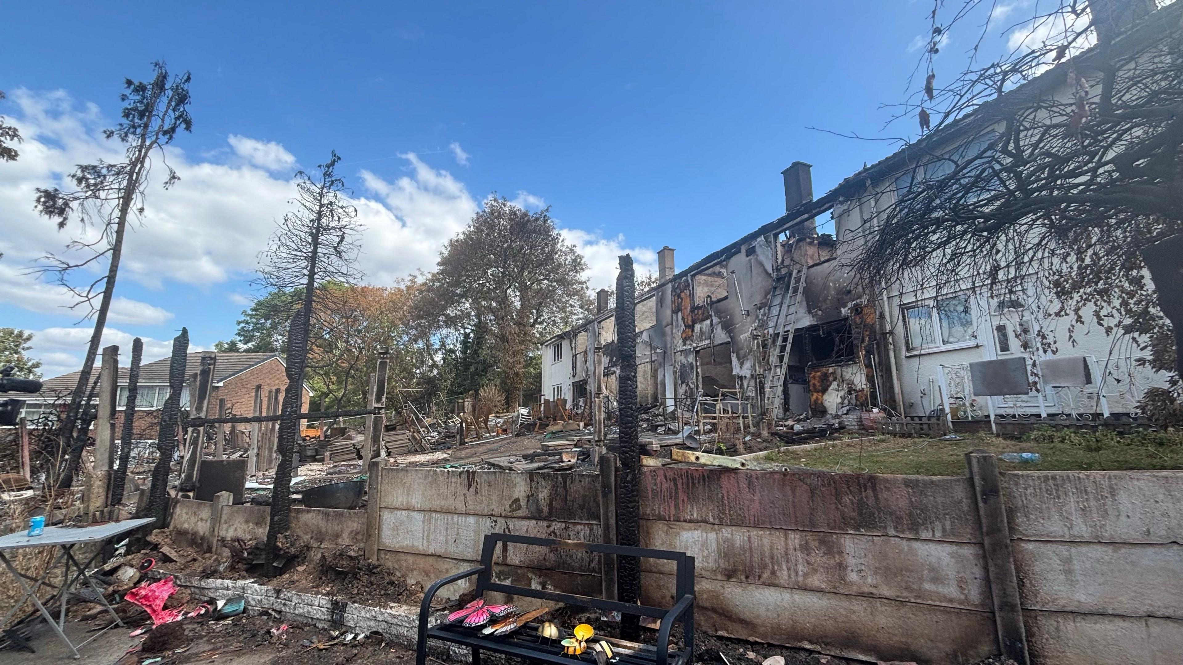 A row of residential properties is shown after a major fire, with extensive damage visible across several homes. Charred walls, burnt-out rooms and blackened rooflines can be seen, while the foreground contains scorched fencing, debris and the remains of trees or garden structures that have been reduced to blackened posts. A ladder leans against one of the damaged buildings, and scattered household items remain among the wreckage. The scene is photographed in daylight under a bright blue sky, highlighting the scale of the destruction.