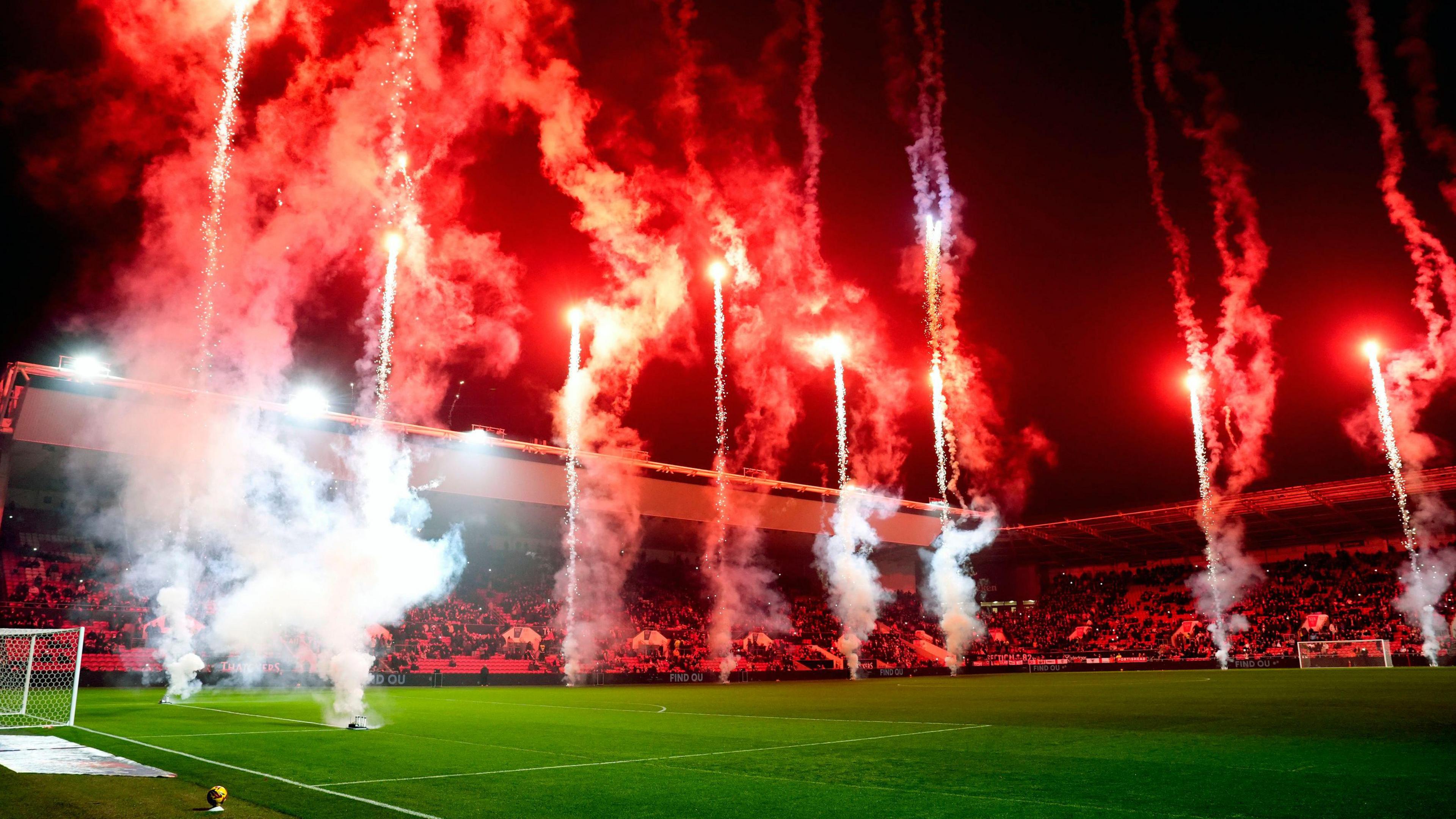 People sitting in Ashton Gate Stadium whilst red fireworks are let off from the pitch