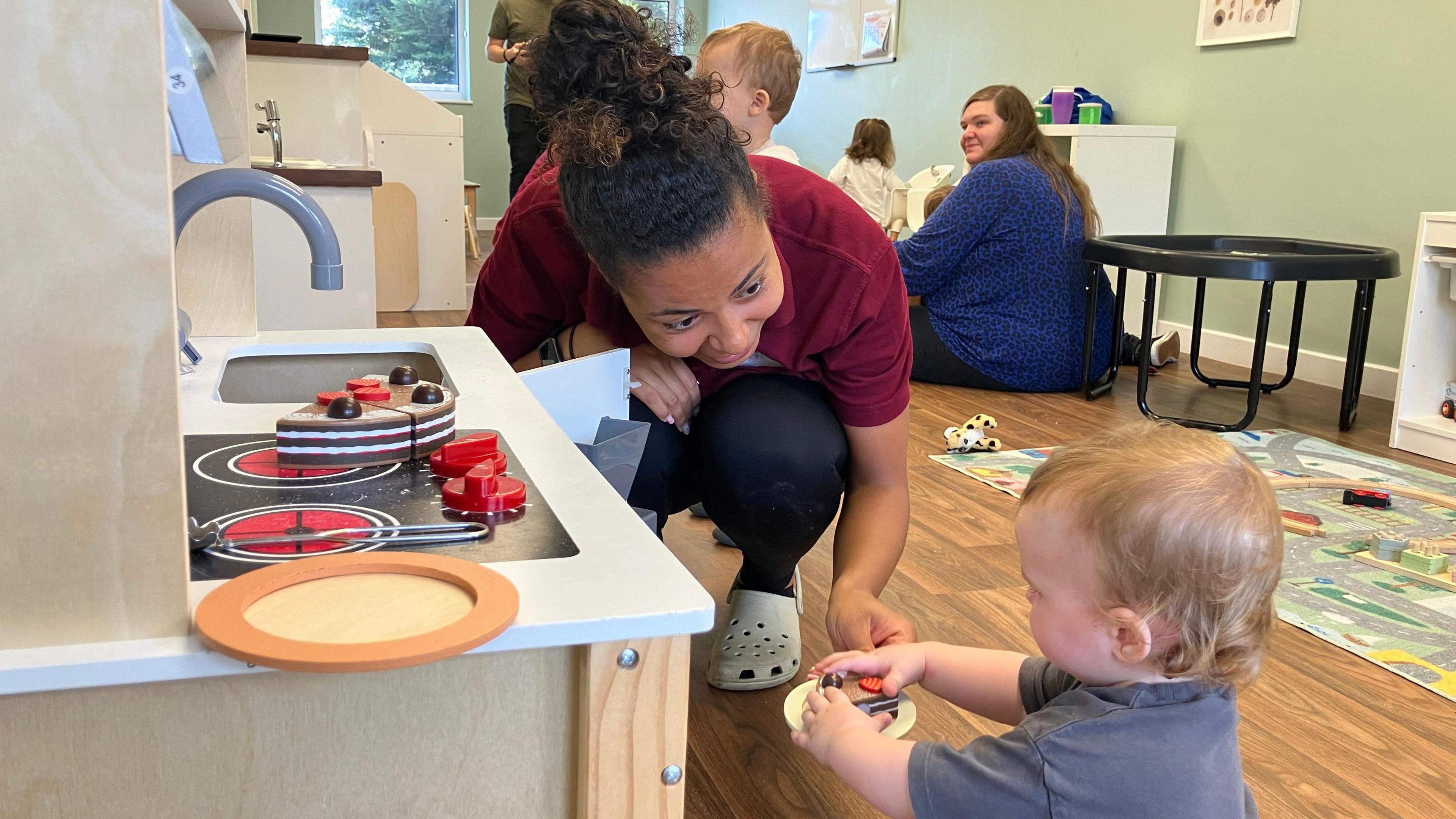 A nursery worker crouches down to the floor to play with a child at a kitchen play-set. The woman, who has her curly black hair tied up, is smiling at the child who is playing with a toy plate.