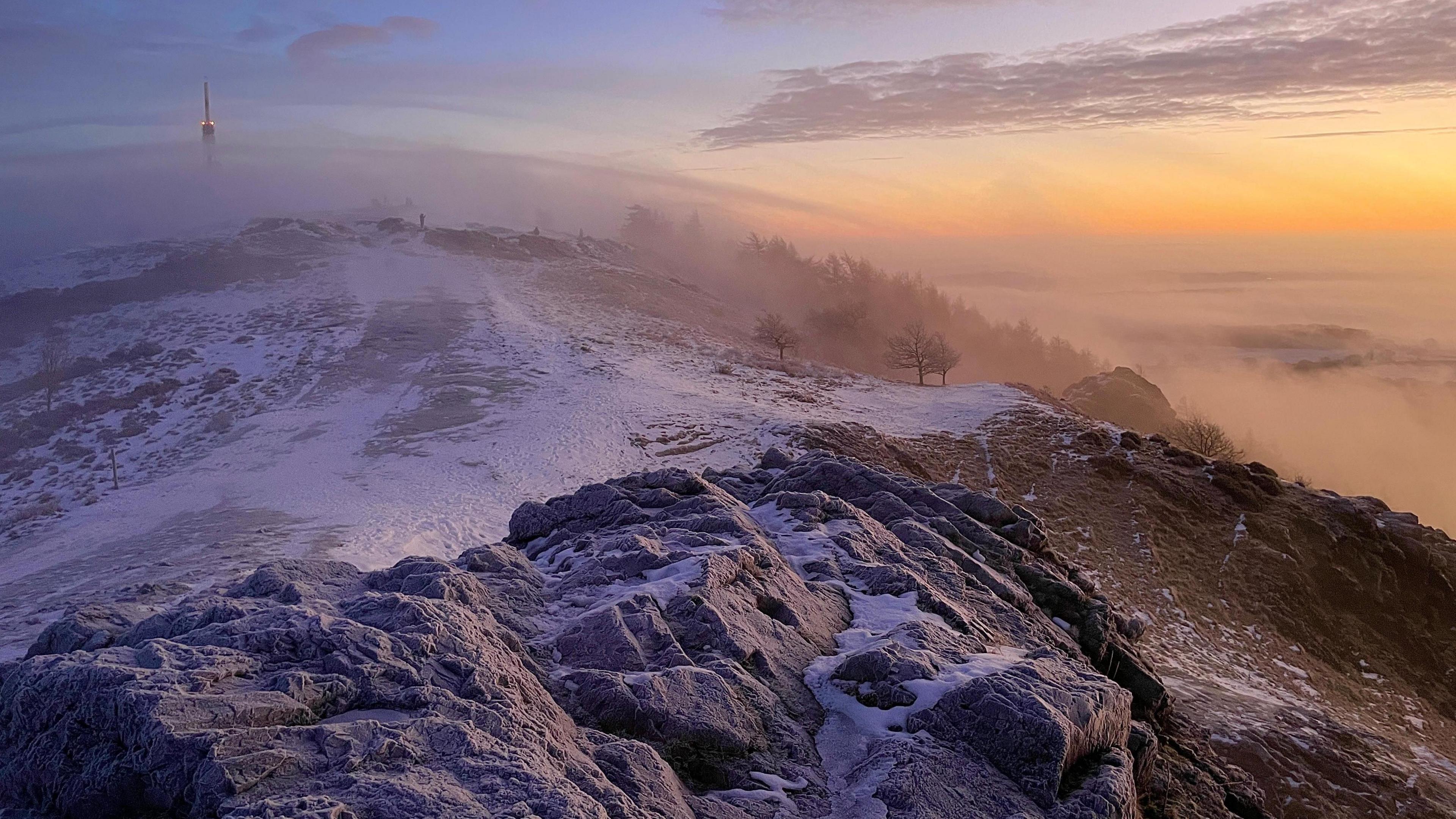 The top of a hill is covered in snow and grey rocks. There is a sunrise to the right of the picture and a fog spreads throughout the sky.