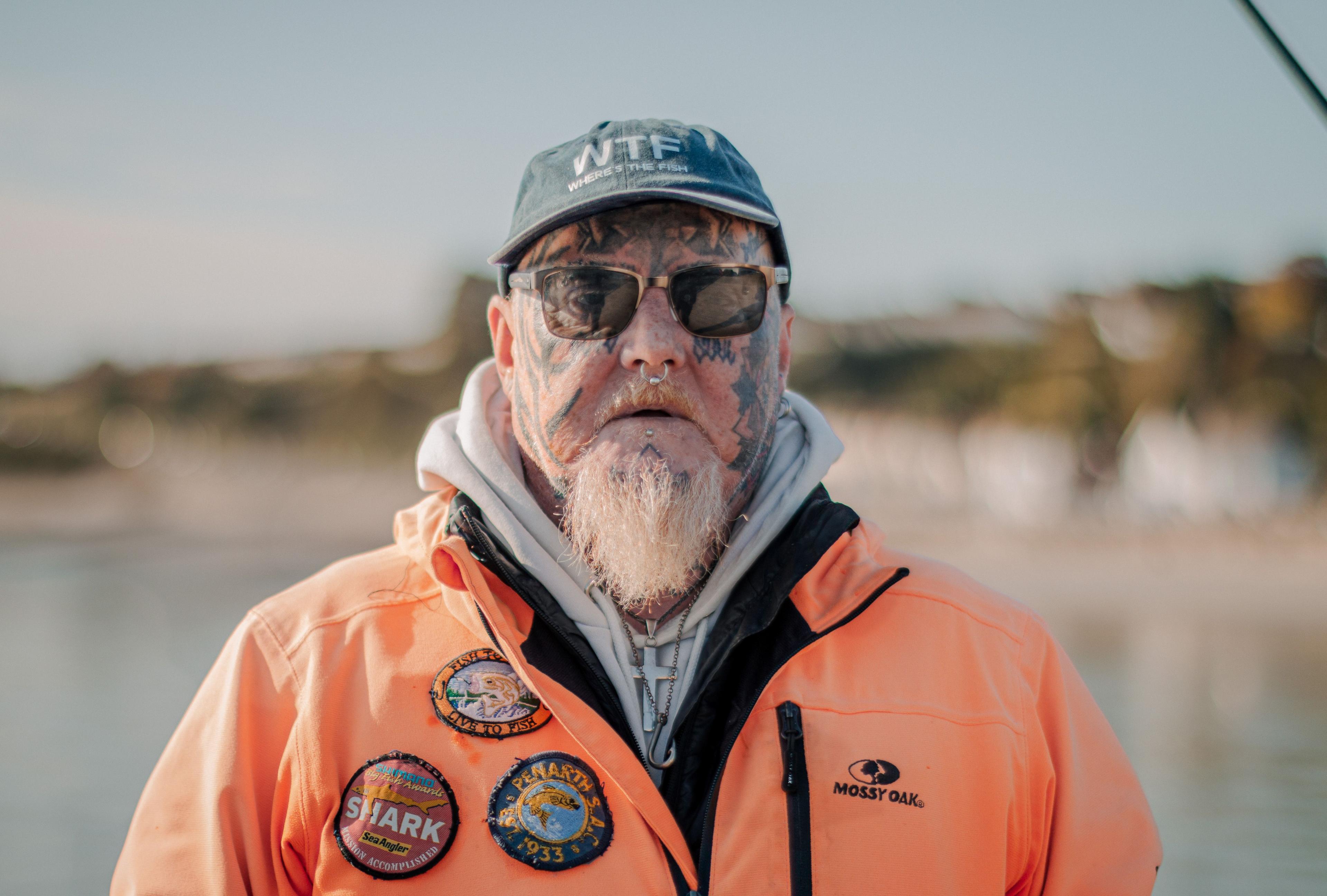 Angler Jay Lewis who has a white beard, facial tattoos and a septum nose ring is wearing a grey cap, orange waterproof jacket and is looking at the camera 