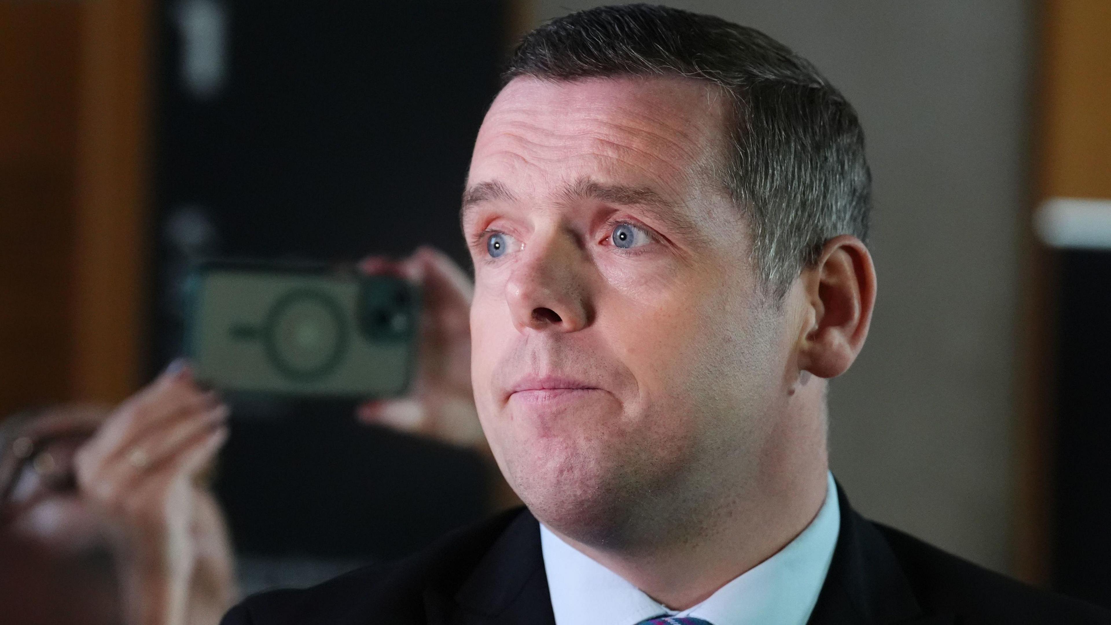 Douglas Ross looking off camera. He has dark, greying hair and is wearing a blue shirt.