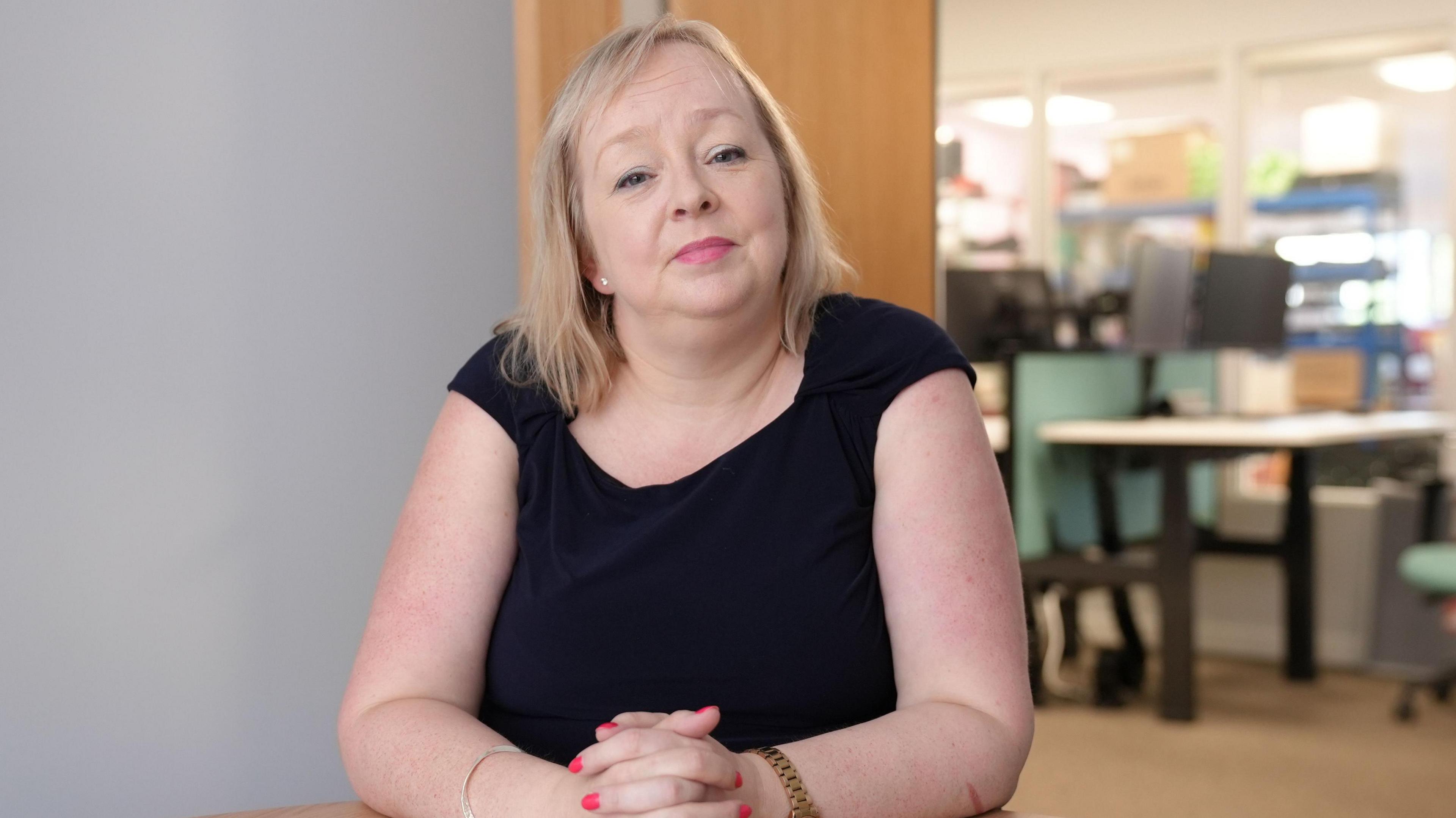 Julia is wearing a dark navy, sleeveless dress. She is sat in her offices, with her hands clasped in front of her, on a desk. She has shoulder length, straight blonde hair. Behind her is an open door to the office, which is slightly blurred.