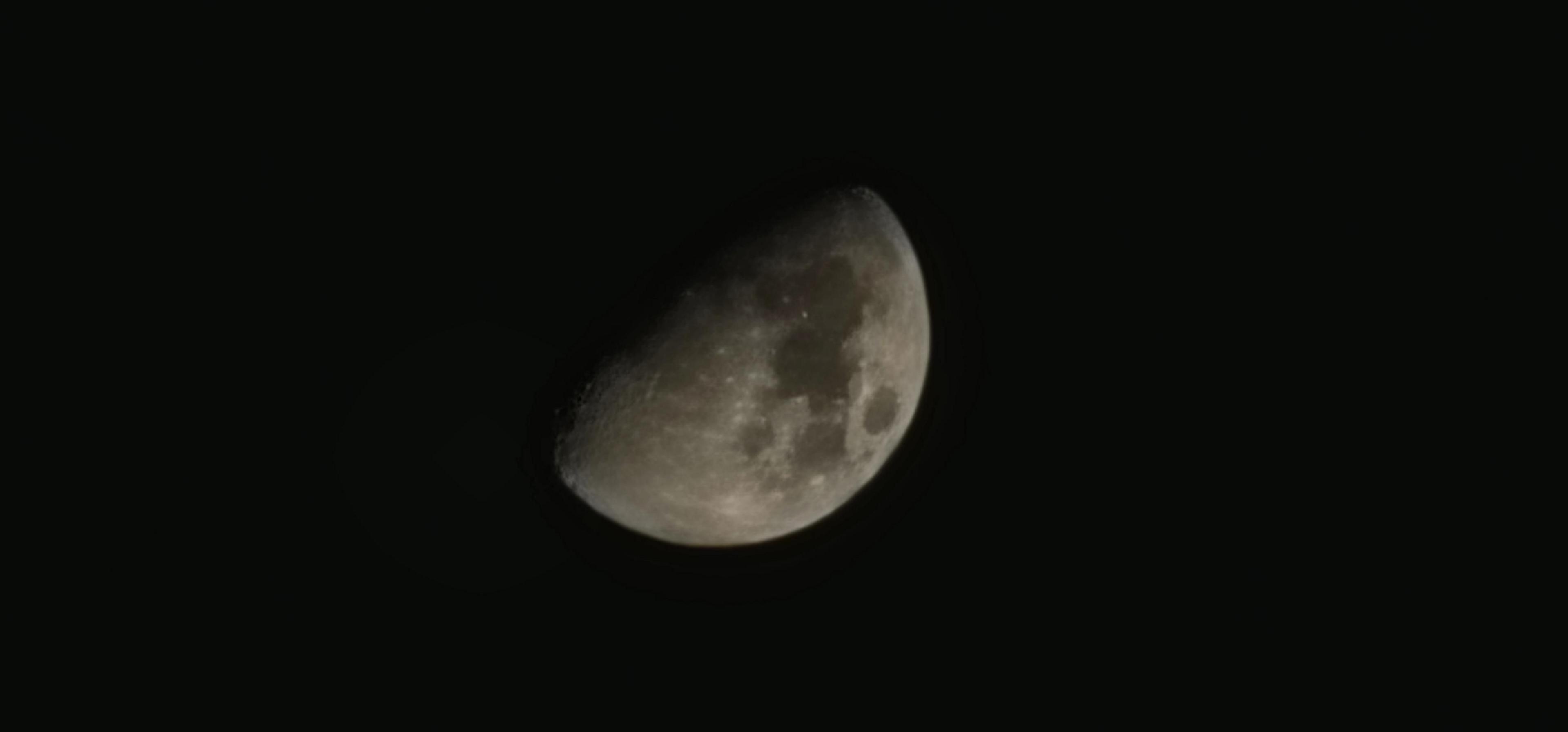 The moon in a black sky, showing several features on its rough landscape. About half of the moon is showing with the remainder disappearing into shade.