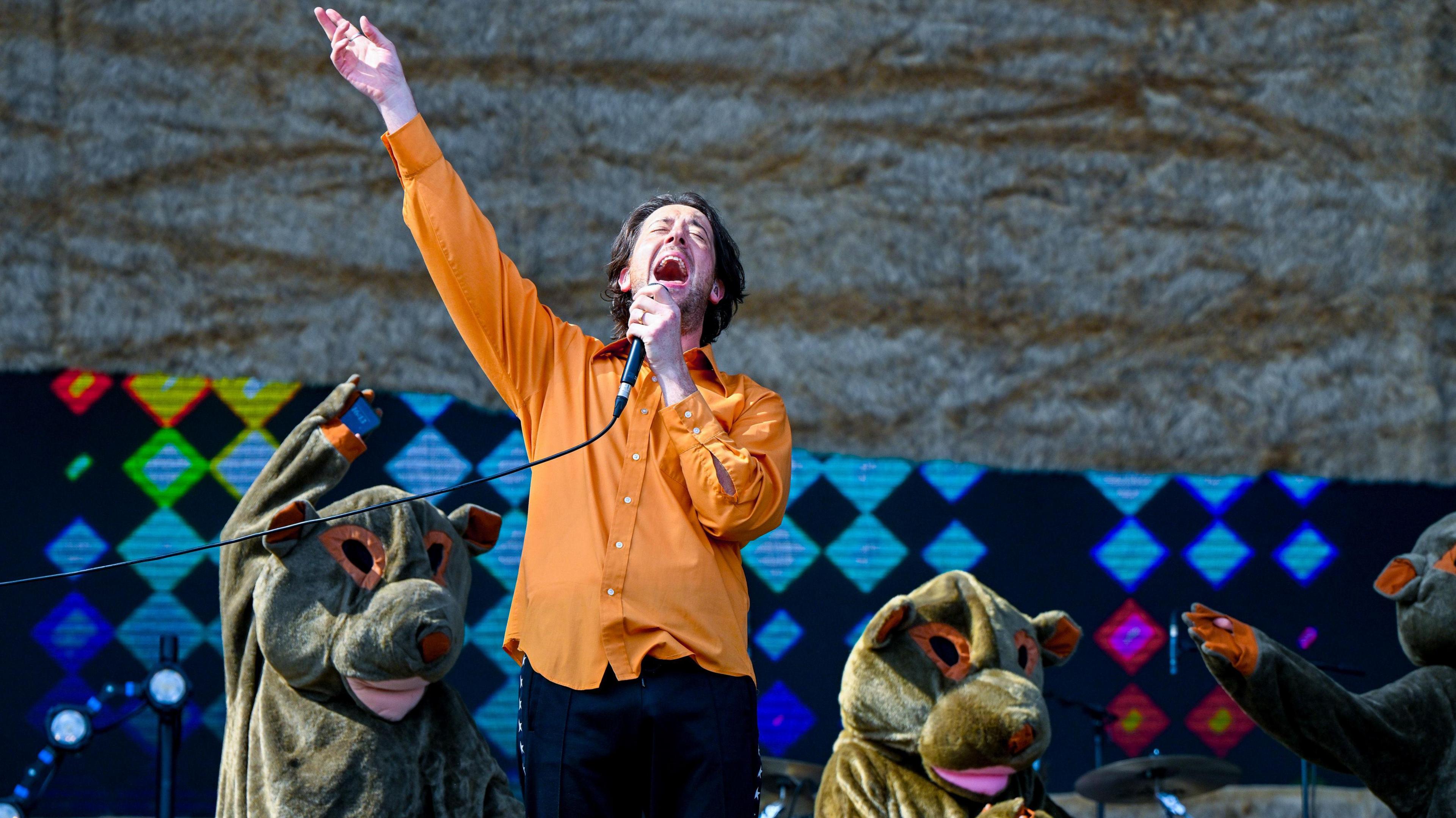 Matthew Murphy of The Wombats sings into a microphone on stage. Behind him are people dressed as animals, perhaps wombats?