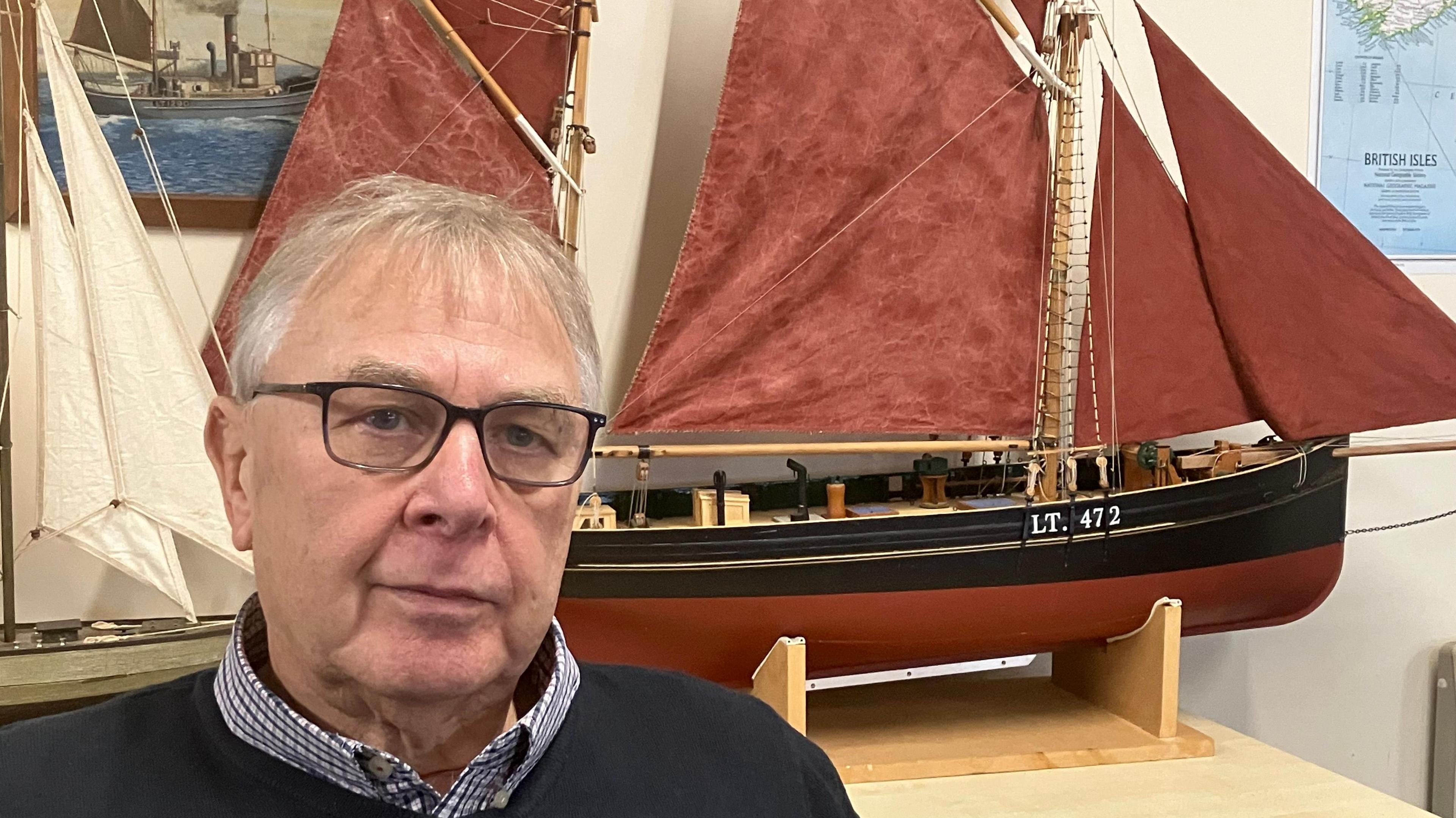 Gerry Skews wearing a blue jumper, checked shirt and glasses sits in front of a model of the Excelsior, a Lowestoft-built sailing smack with a large red sail