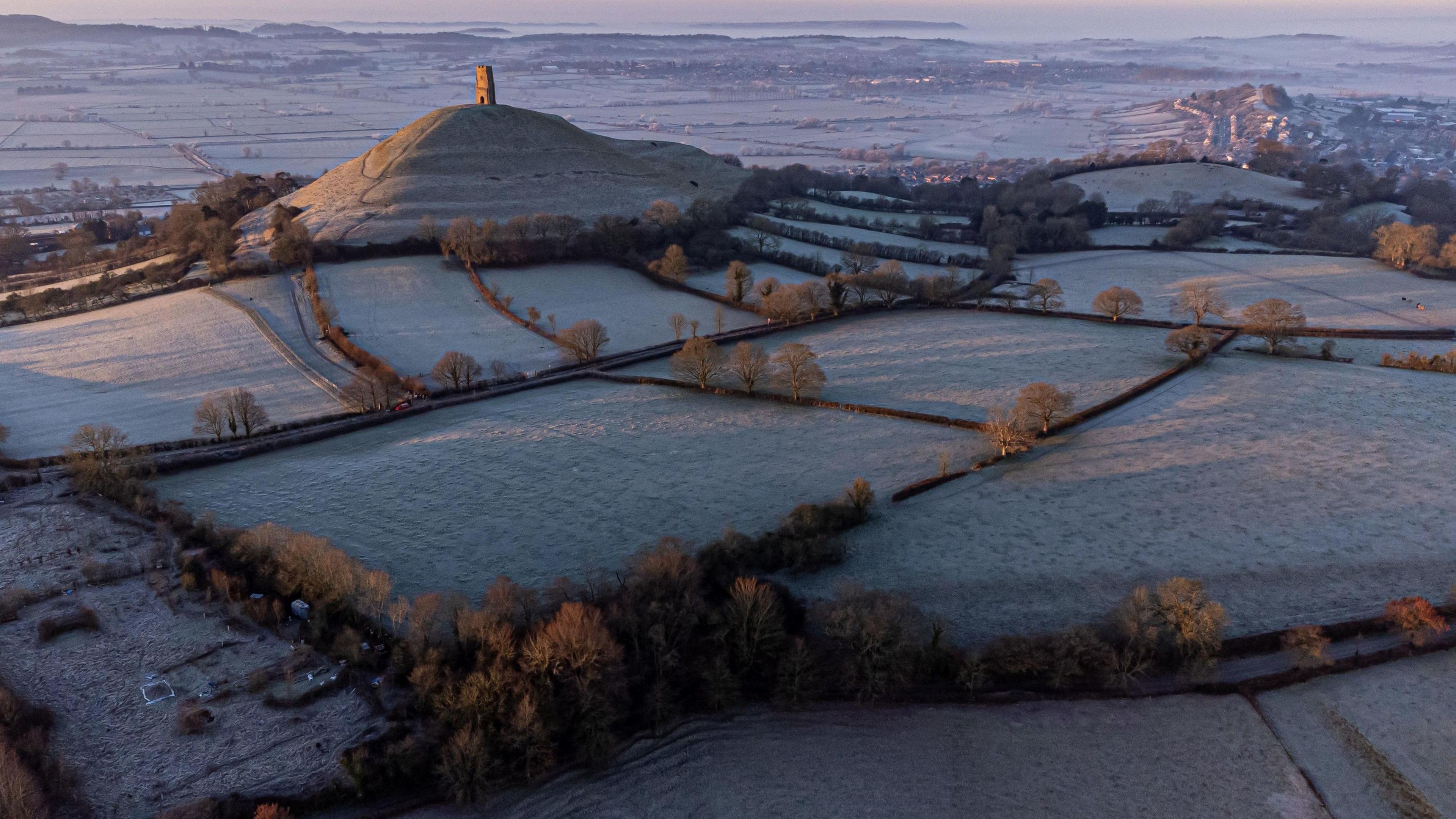 A wide shot of the Somerset Levels at dawn with Glastonbury Tor in the centre and fields around it covered in frost