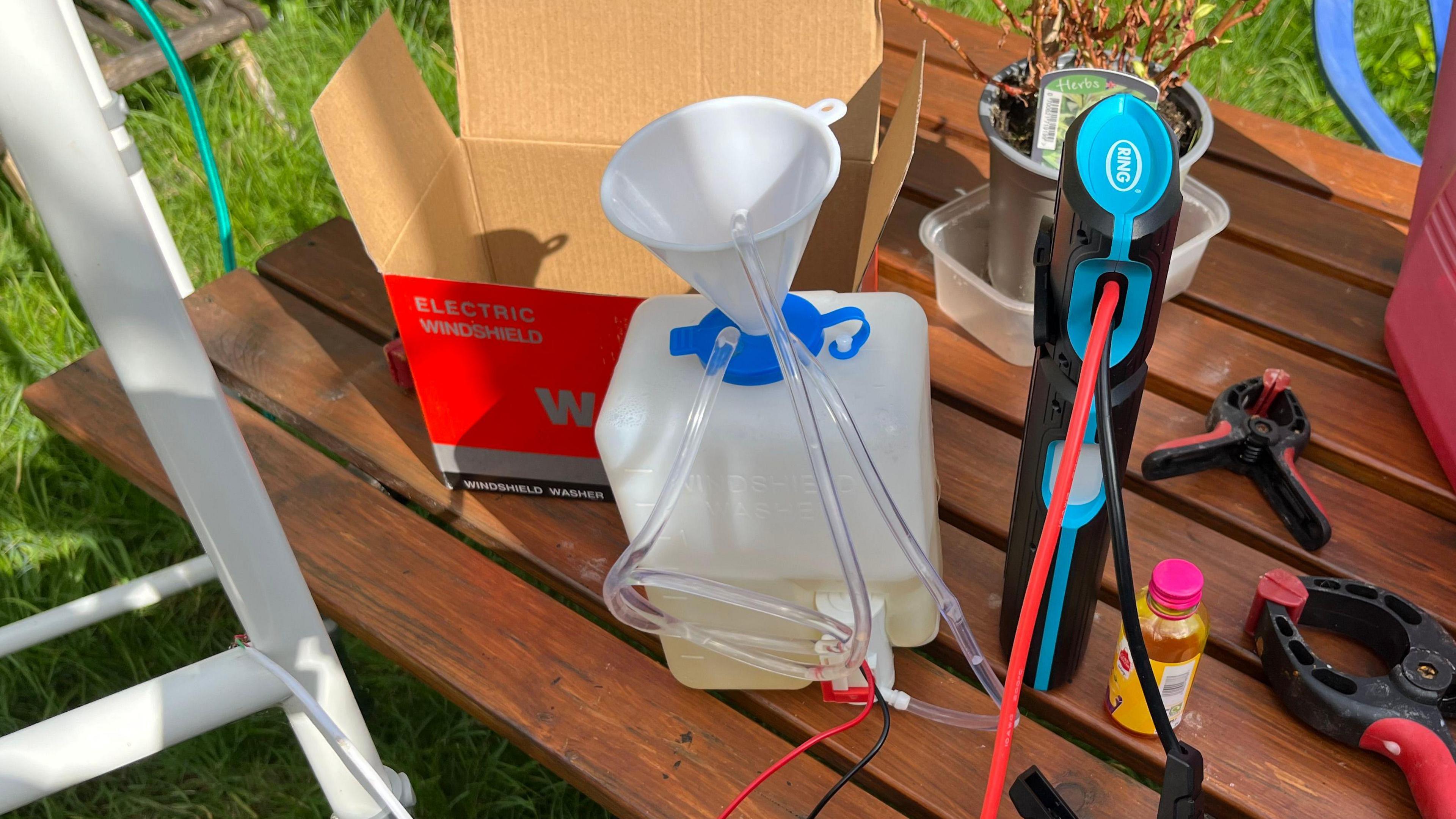 A water tank reservoir from a car holds about 1.5 litres of liquid, there is a large battery with various tubes and wires emerging from it and the bottle. It is all on a garden bench.