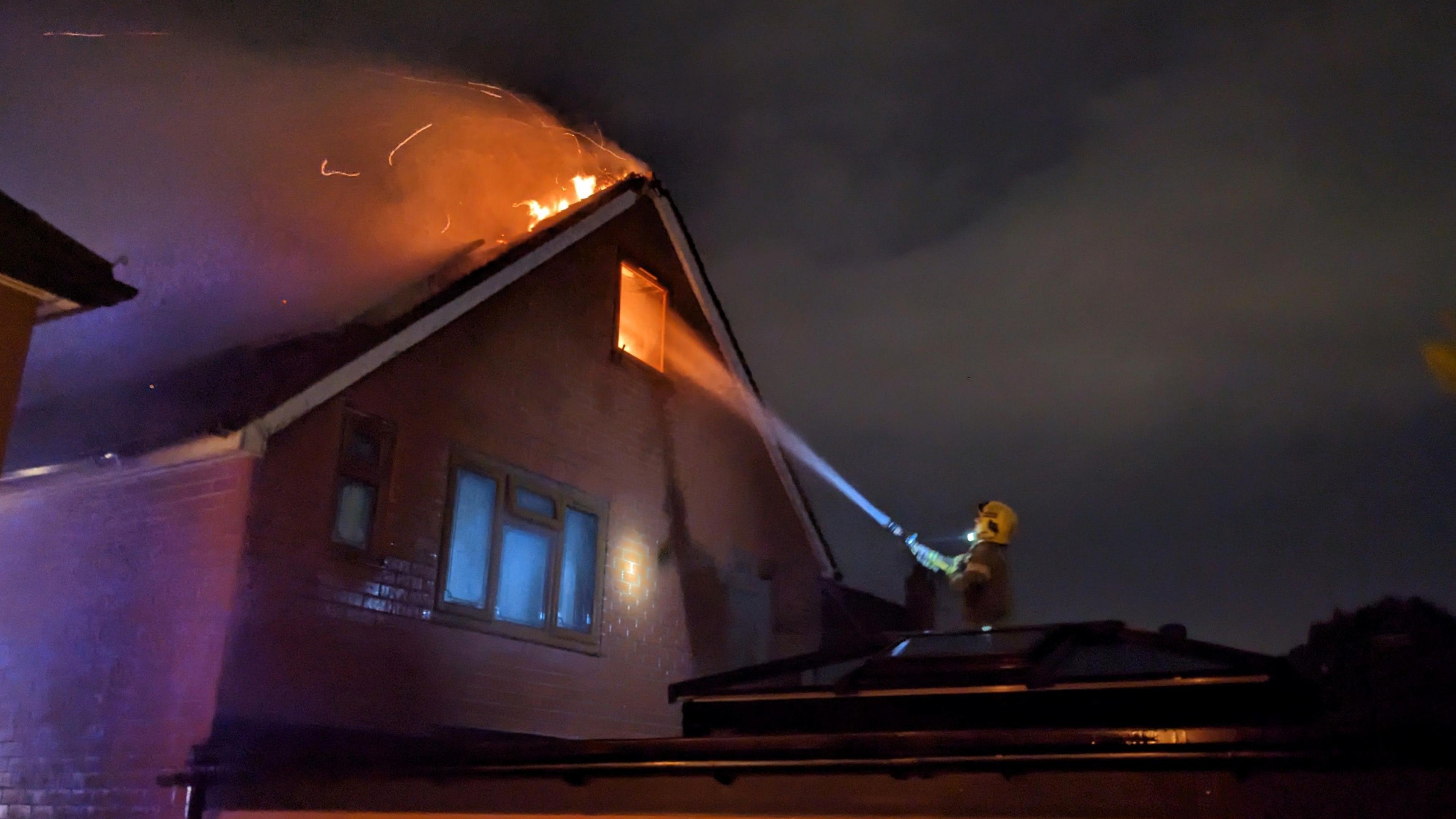 A house, at night, with its roof on fire and a firefighter spraying water on the flames with a hose jet.