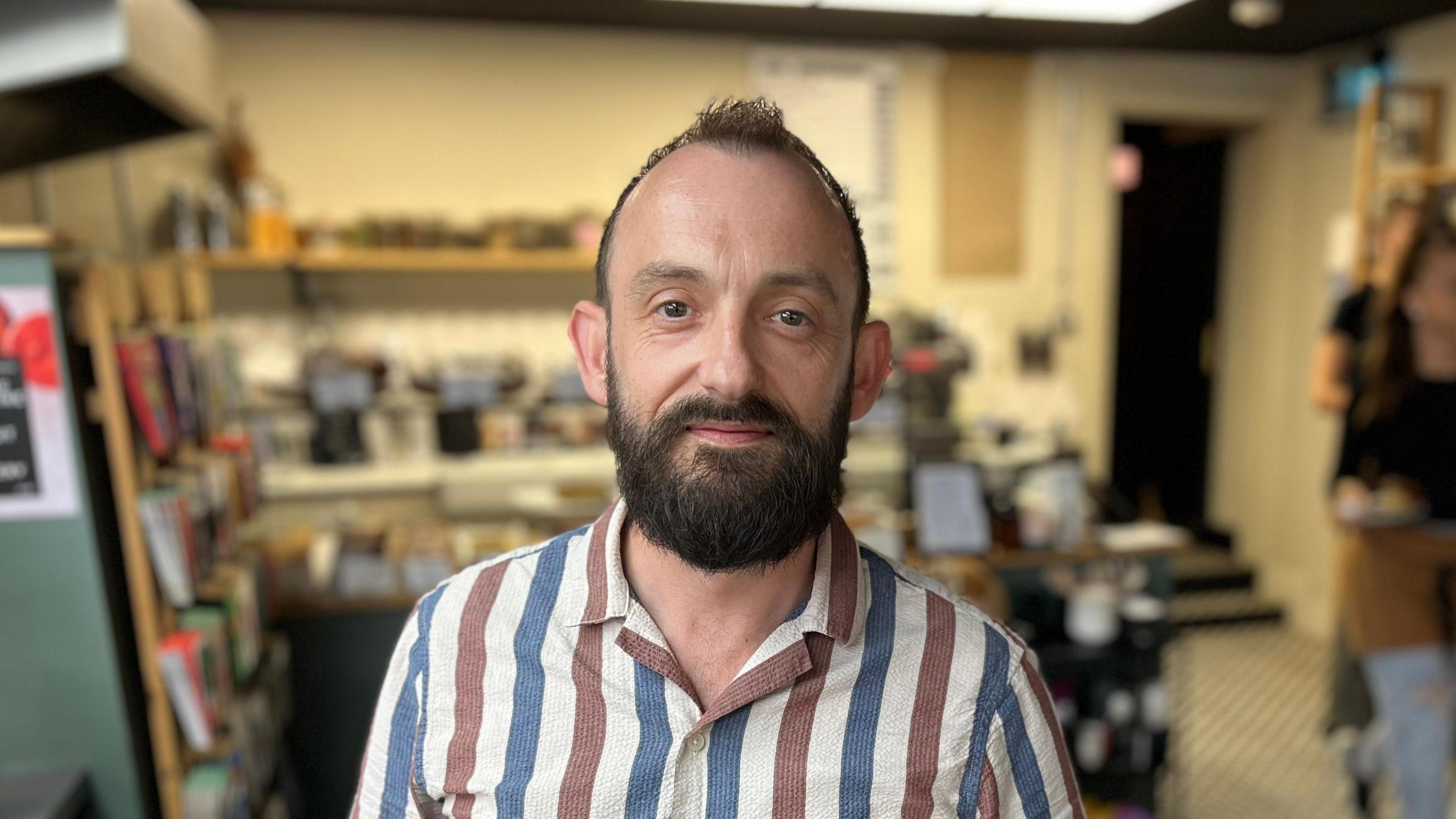 Alex Middleton from the shoulders up. He has brown hair and a brown beard. Behind him is the cafe out of focus.