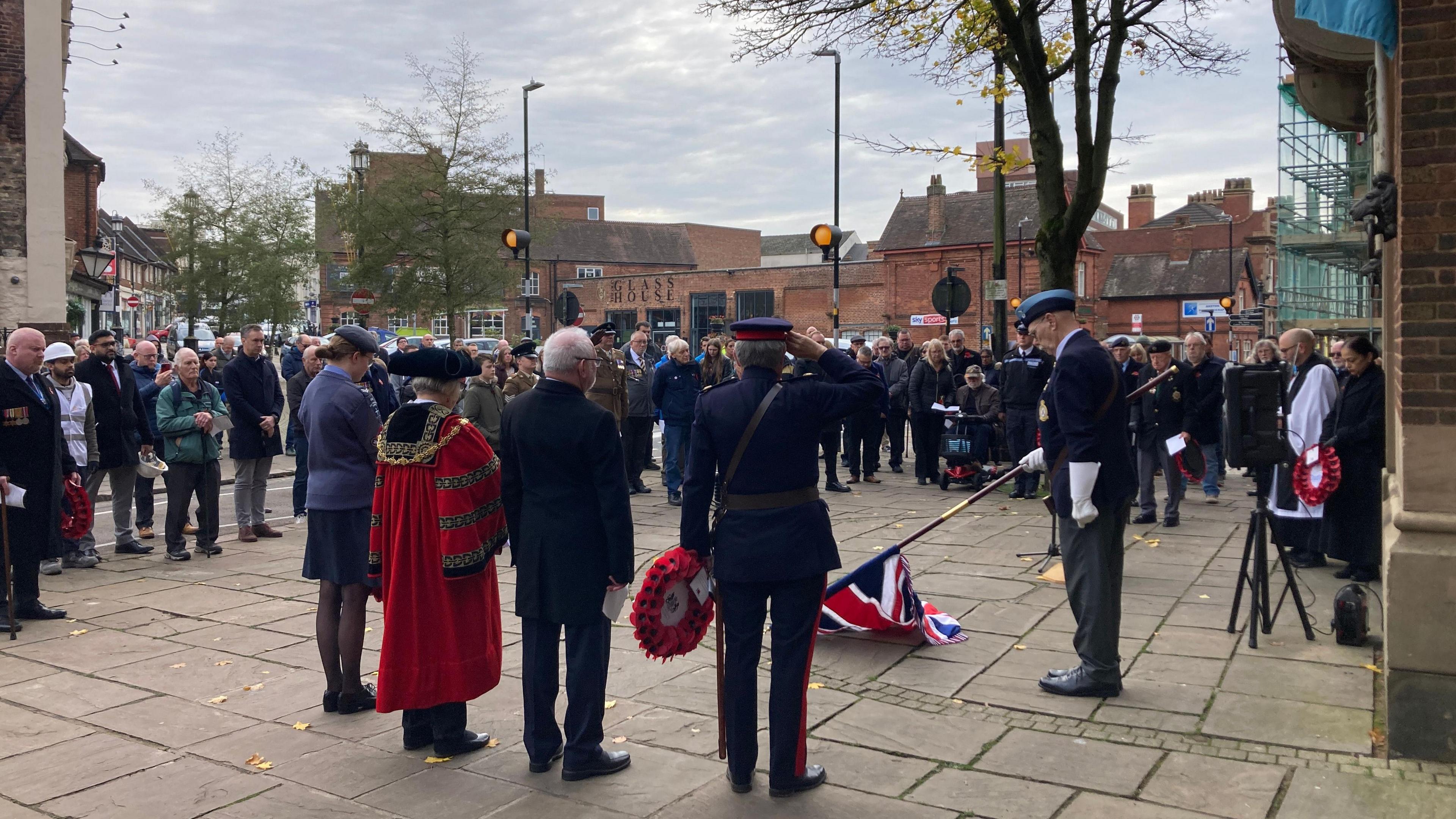 Representatives gather with members of the public to observe a moment's silence. One man is holding a poppy wreath while another is holding a flag. 