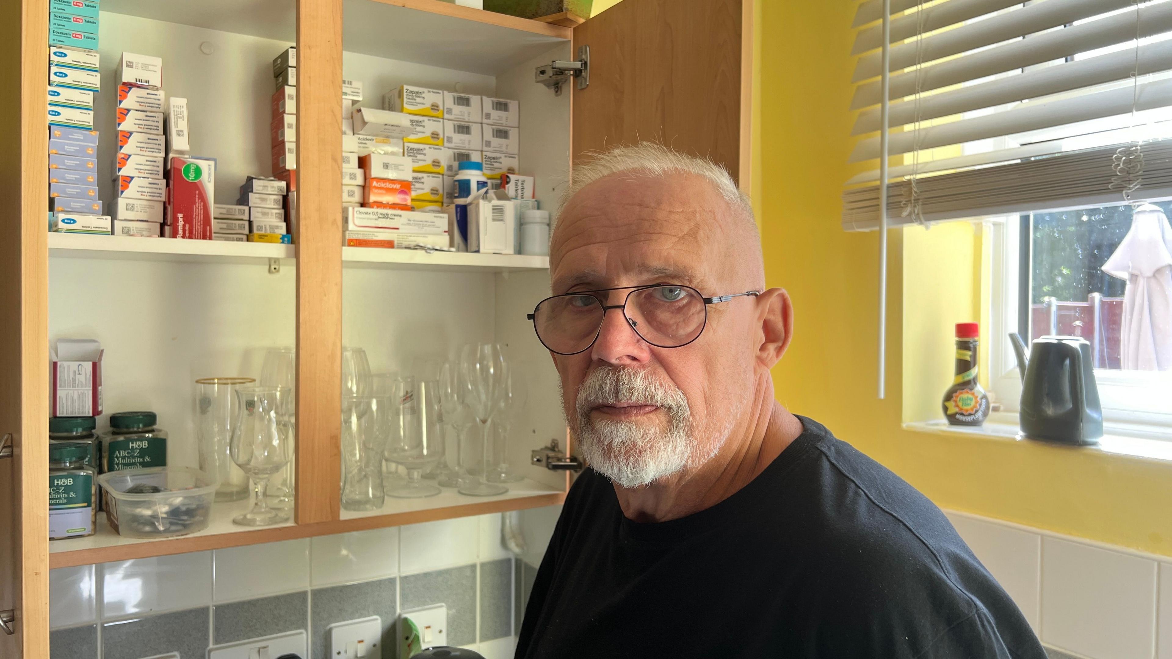 69 year old man with a white goaty beard, black rimmed glasses and thin white hear wearing a black t-shirt looking at the camera in his kitchen.  A cupboard is open displaying a large number of medicines.