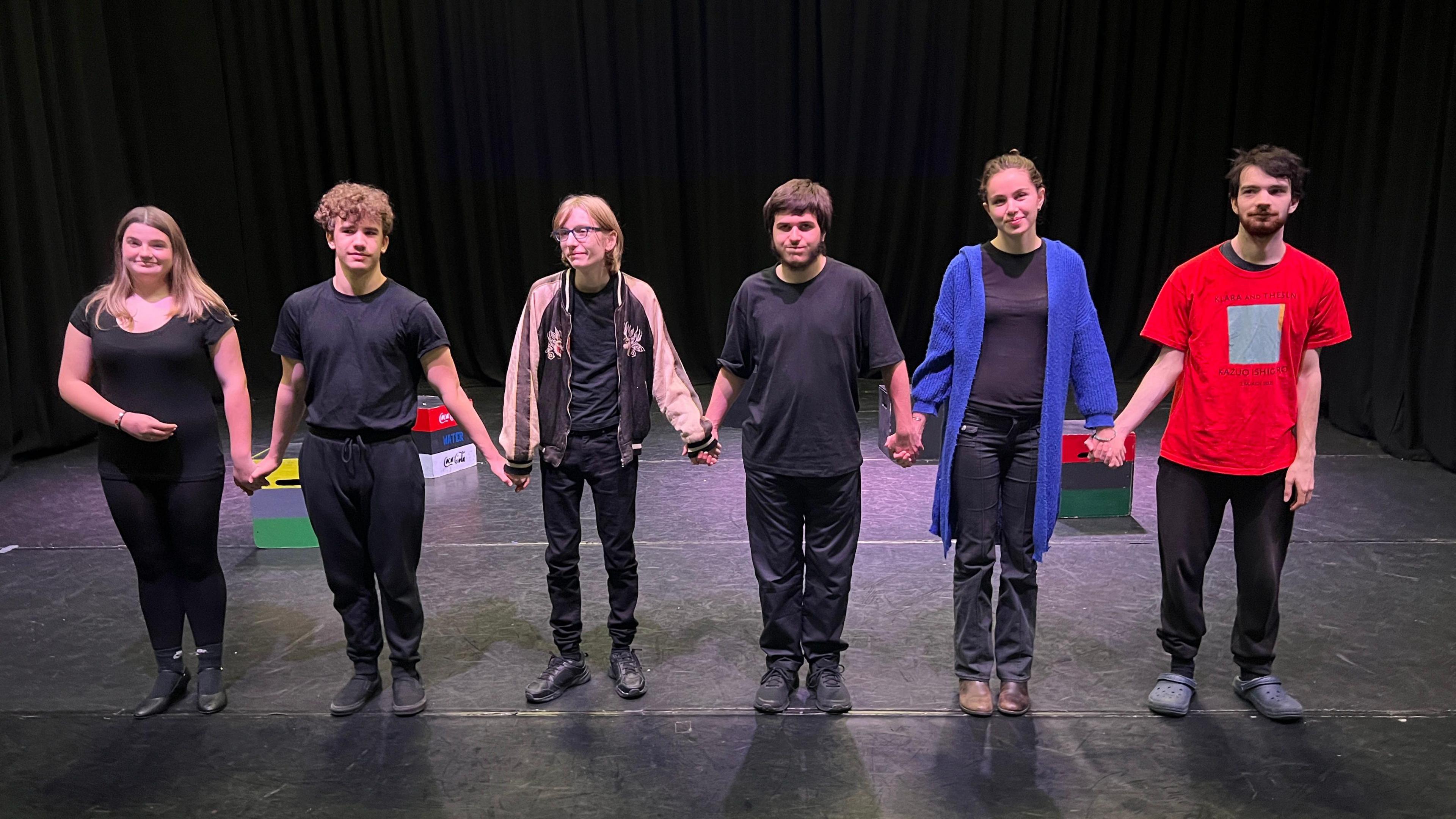 Six people, most dressed in dark T-shirts and trousers, holding hands on a black stage.