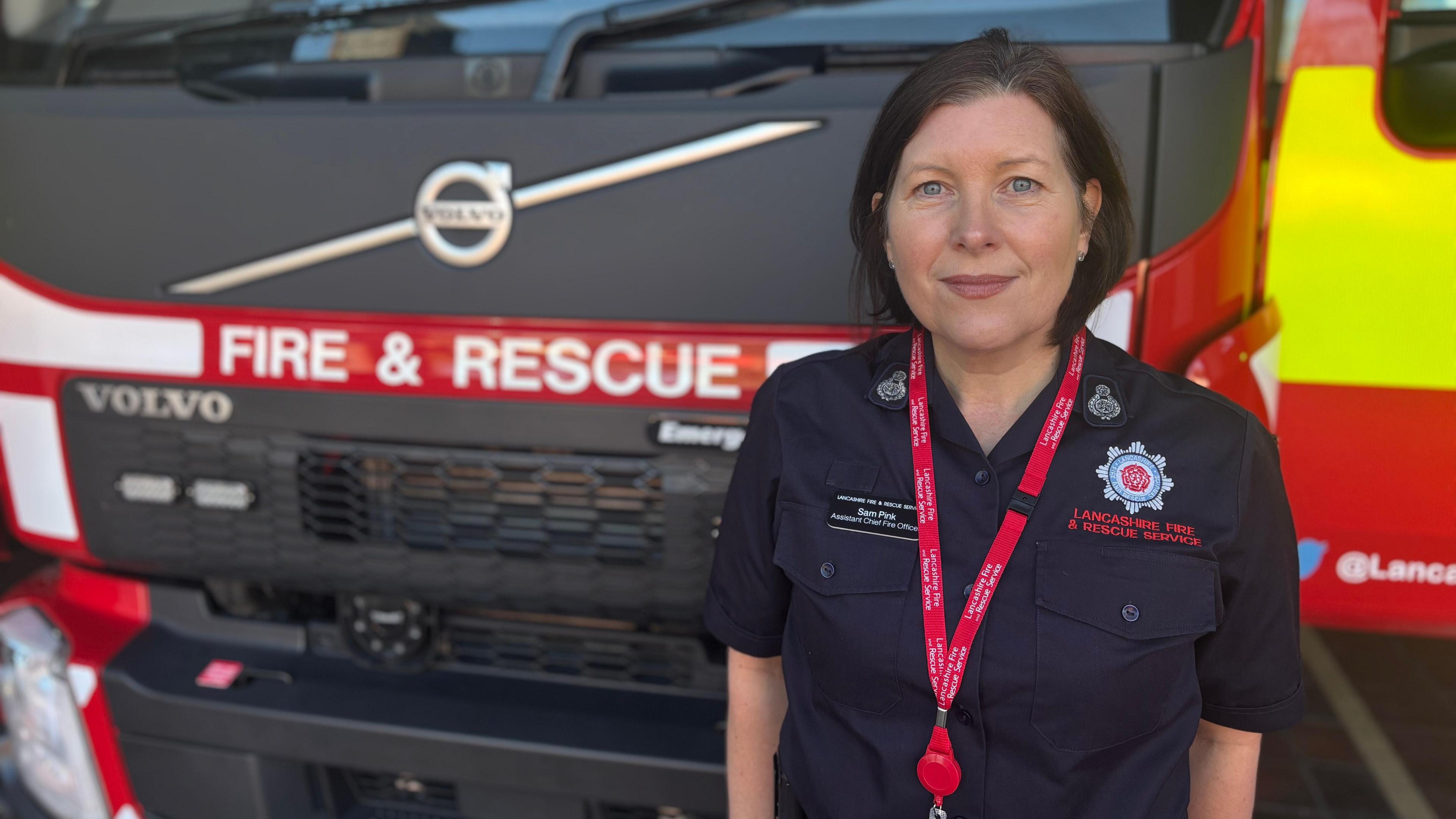 Samantha stands in front of a fire engine. She wears a navy blue Lancashire Fire and Rescue service uniform and a red lanyard. She has bobbed brown hair and smiles at the camera.