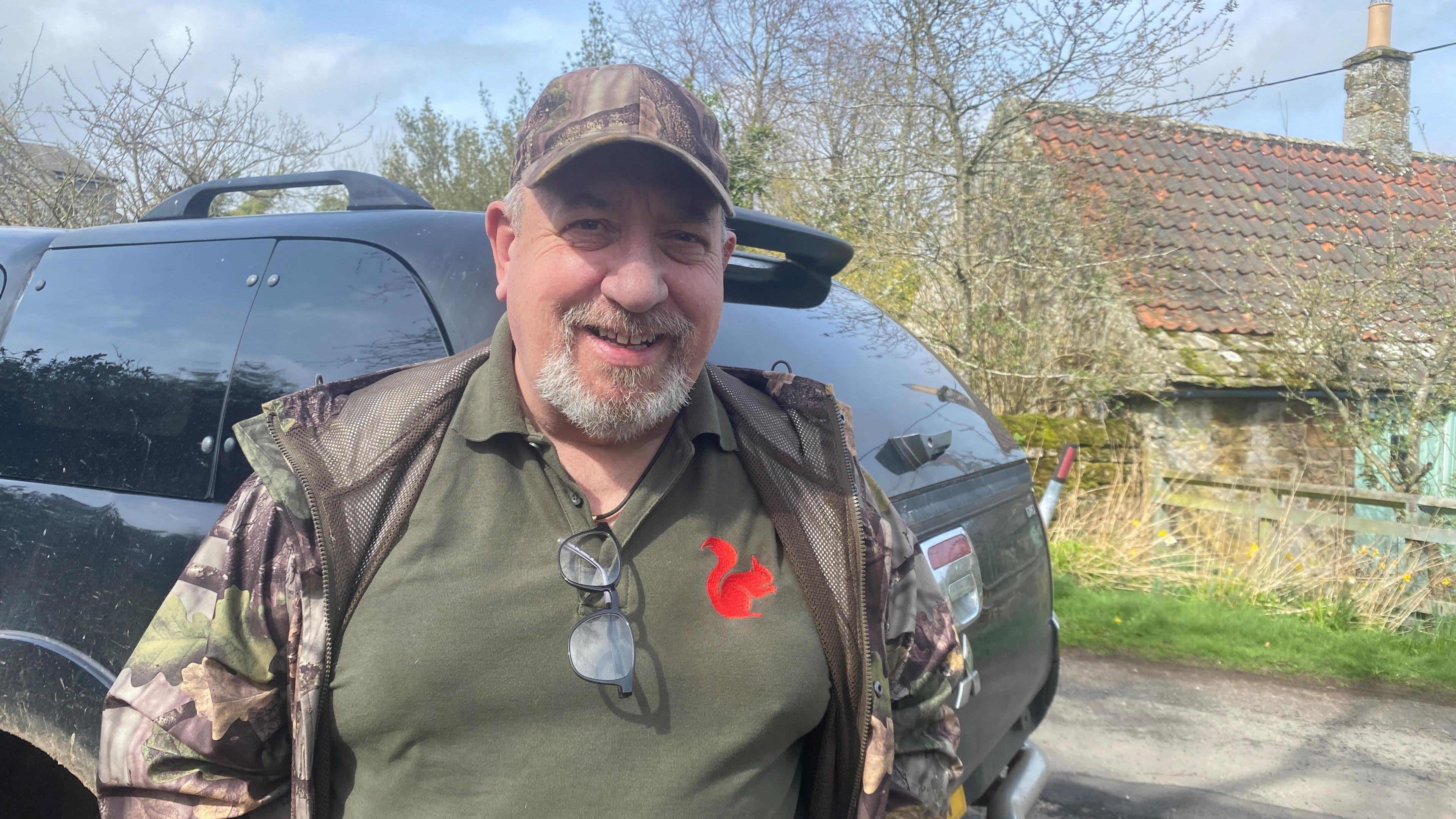 Ian Glendinning is standing in front of a cottage with a black car behind him. He is a man in his 60s with a beard and wearing a green top and jacket and a cap.