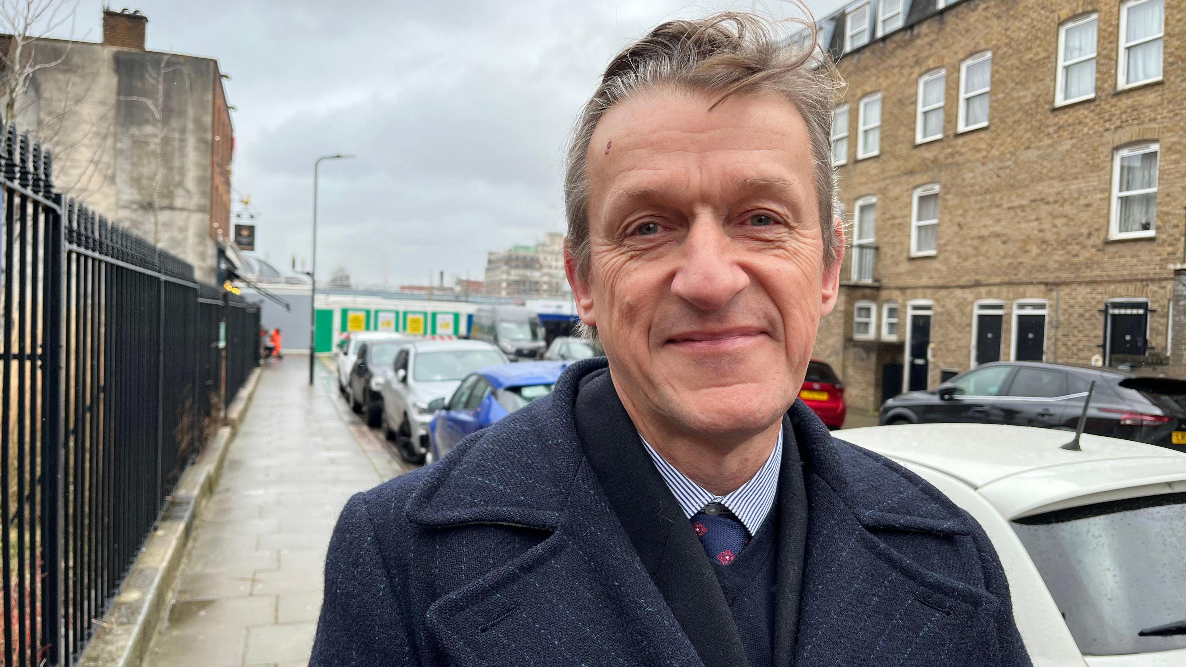 Richard Olszewski wearing a suit with a tie and a navy coat, looks into the camera, smiling. He is standing on a street with the pavement shown on the left and parked cars are on the right.