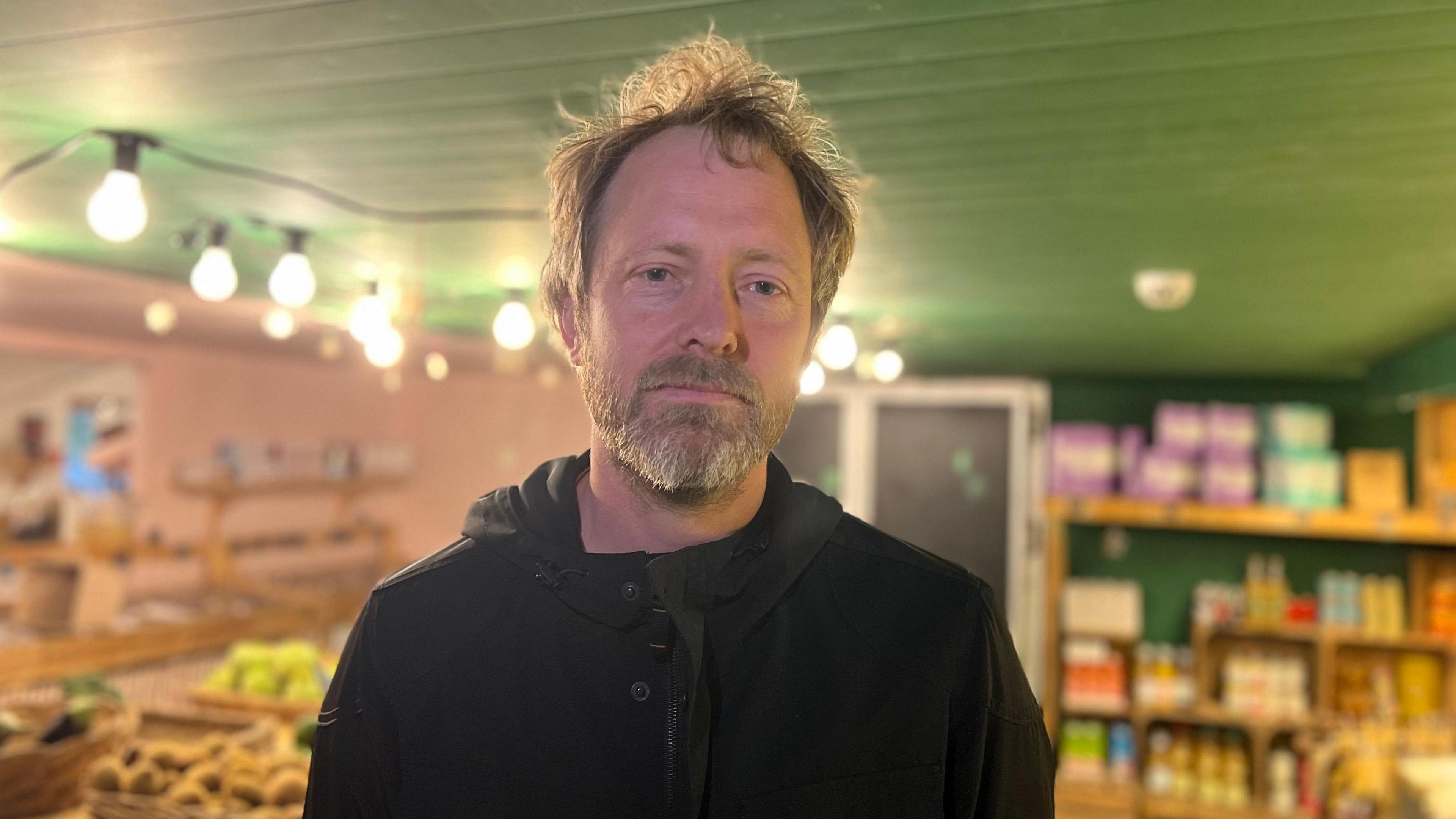 Kaspar Wimberley standing in the SCOOP Sustainable Co-operative store in St Lawrence. He is wearing a black hoodie and has gray hair and a beard.
