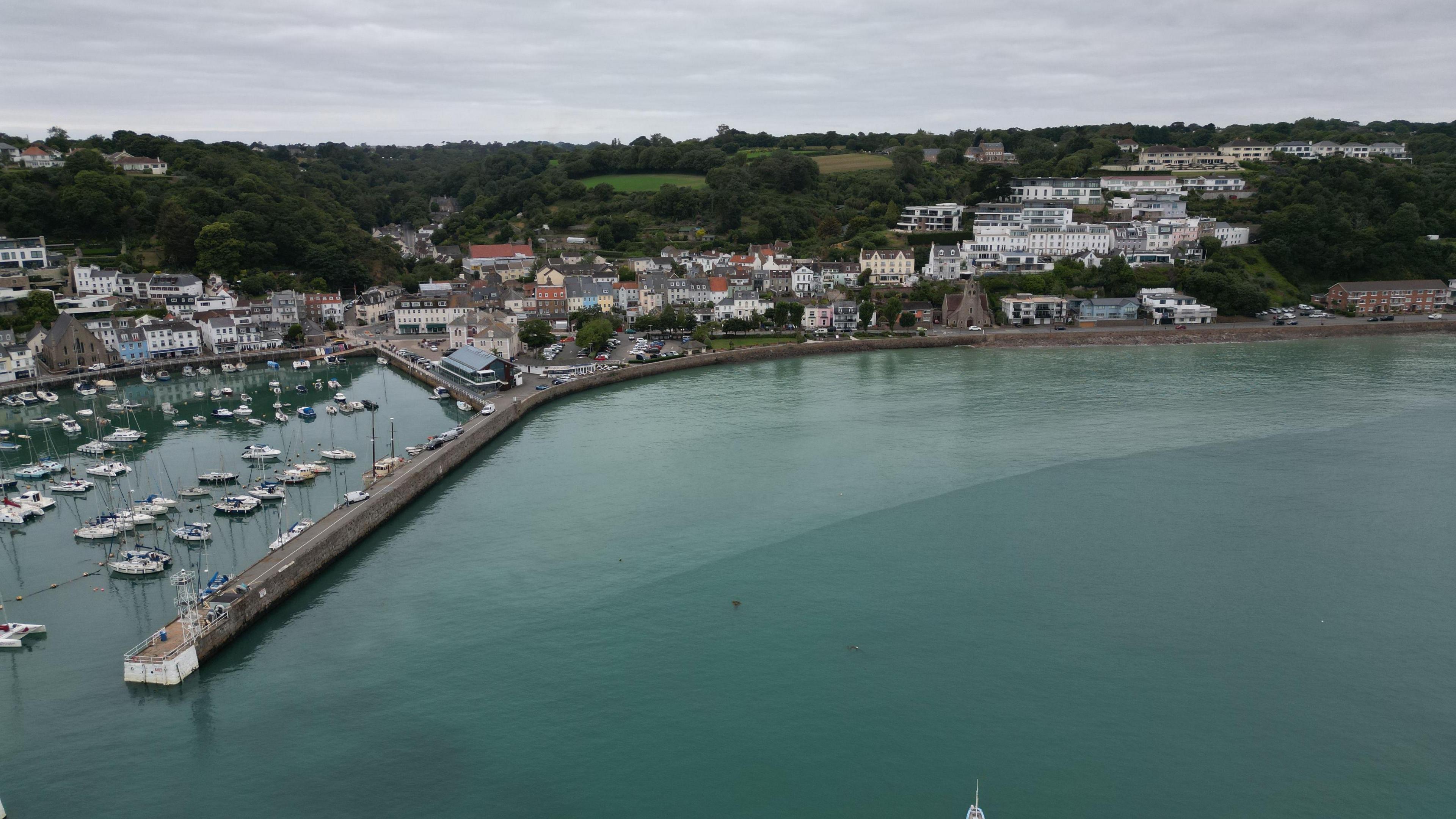 Aerial shot of a harbour.  A pier stretches out into the water. Boats are moored. Houses are on the water's edge. Green trees and fields are on the horizon.