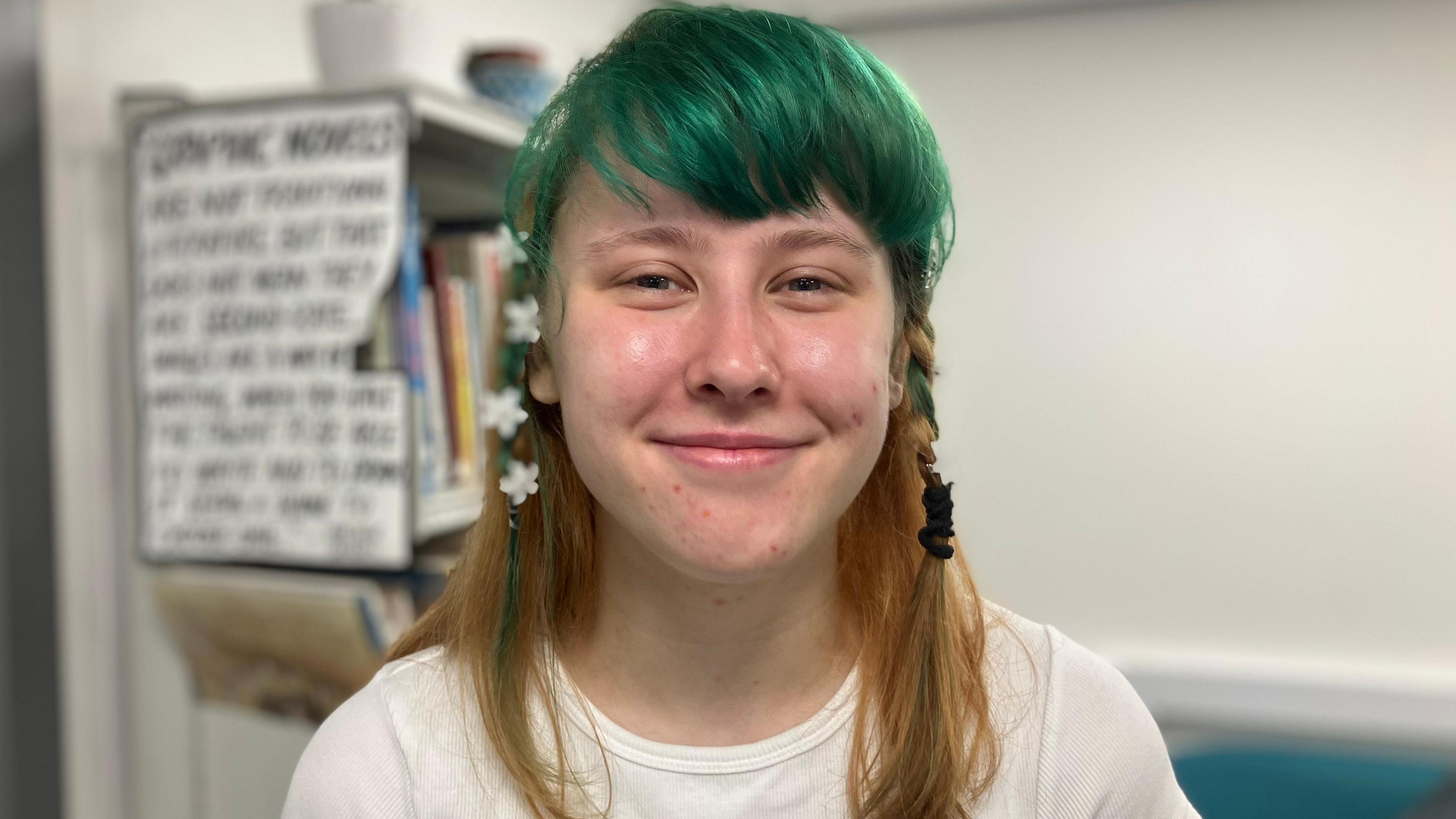 A girl with a green fringe and shoulder length light brown hair, wearing a white t-shirt and standing in a library.