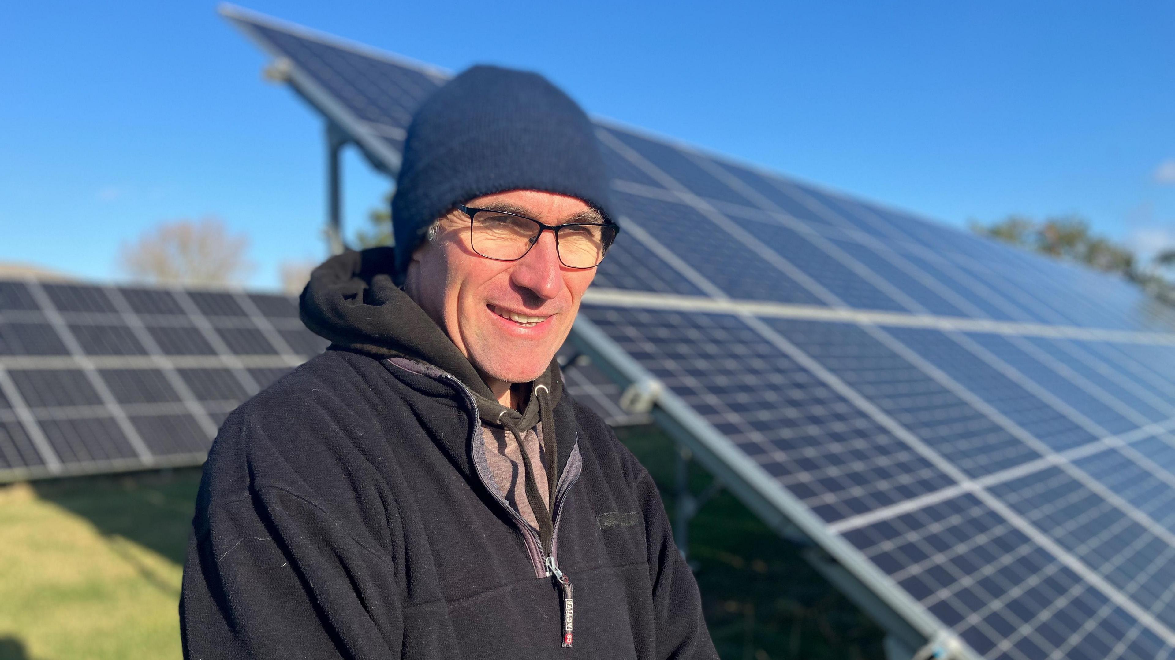 A man wearing glasses and a blue beanie type hat looks at the camera. He is wearing a black hoodie top. Over his shoulder are ground-mounted solar panels.