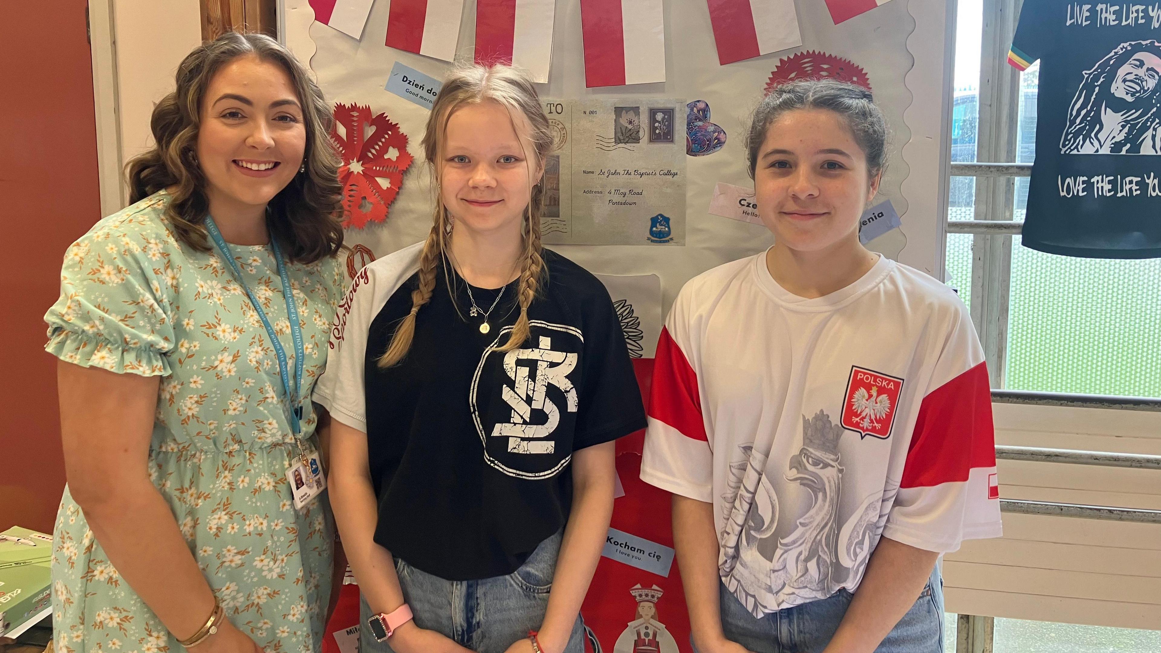 Three girls standing in front of a display for Poland. The girl on the left has light brown hair and wearing a green floral dress and lanyard. The girl in the middle has blonde hair in plaited pigtails and is wearing a black and white t-shirt, necklace and jeans. The girl on the right has brown hair tied back and is wearing a red and white graphic t shirt with Polish tag, and jeans.