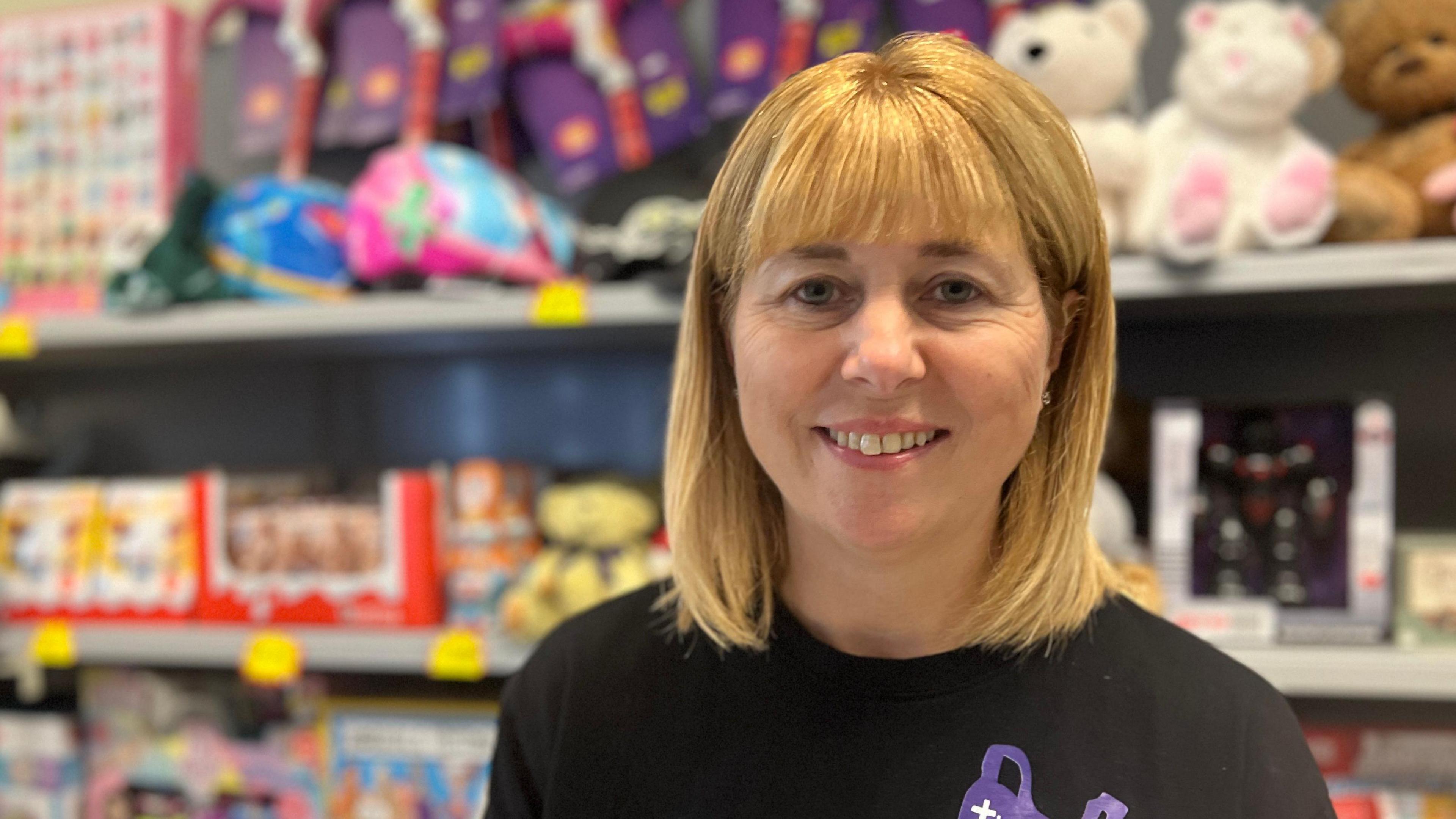 Becky is looking into the camera. She has blonde hair in a shoulder-length bob. She's wearing a black T-shirt with a purple shopping bag logo, and in the background toys and board games can be seen slightly out of focus.