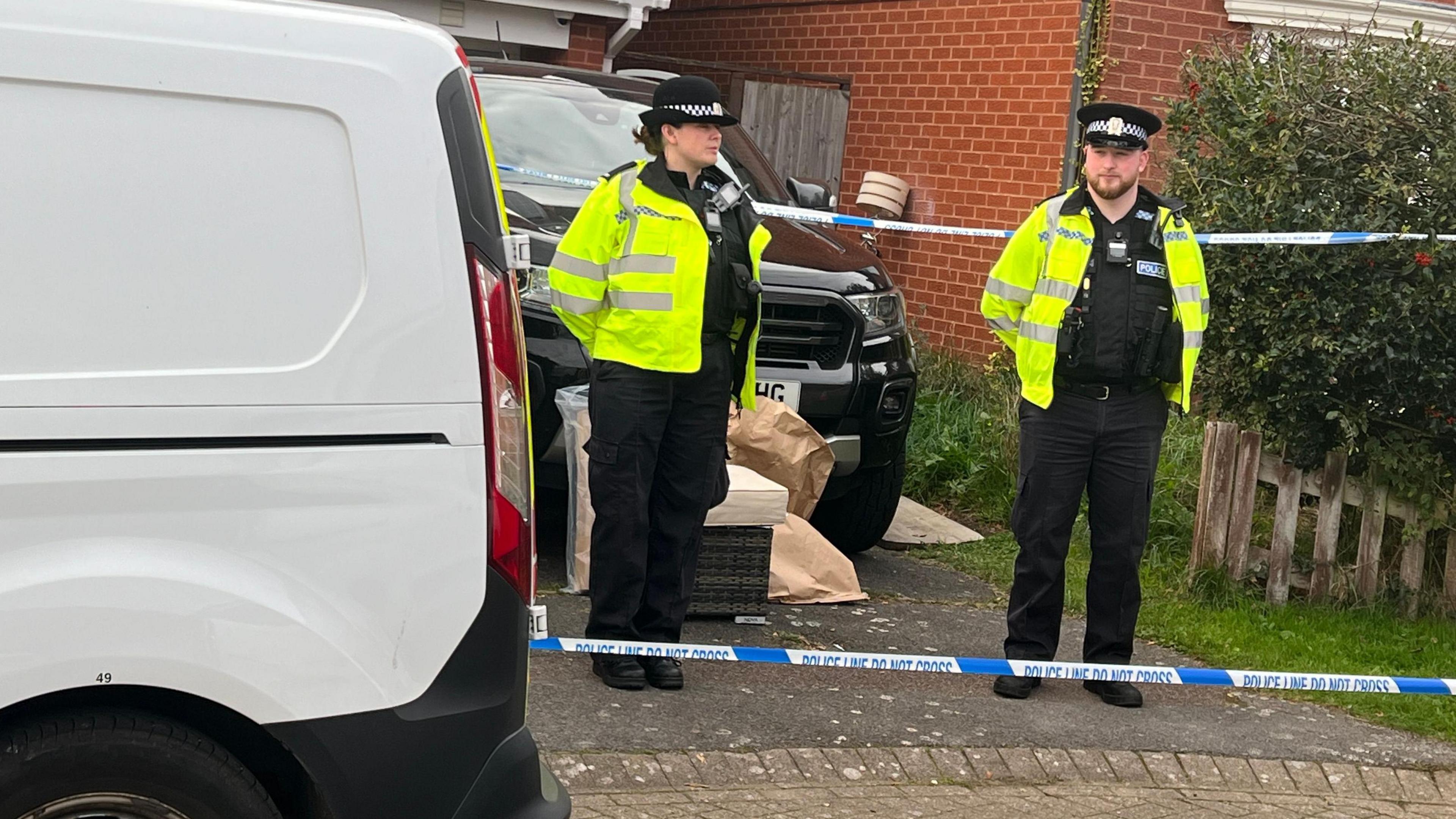 Two police officers stand guard outside a house with police tape stretched across in front and behind them. Some brown bags, a lamp and what looks like garden furniture can be seen in the driveway along with a Range Rover-type car. A white van can be seen parked on the road outside the house.