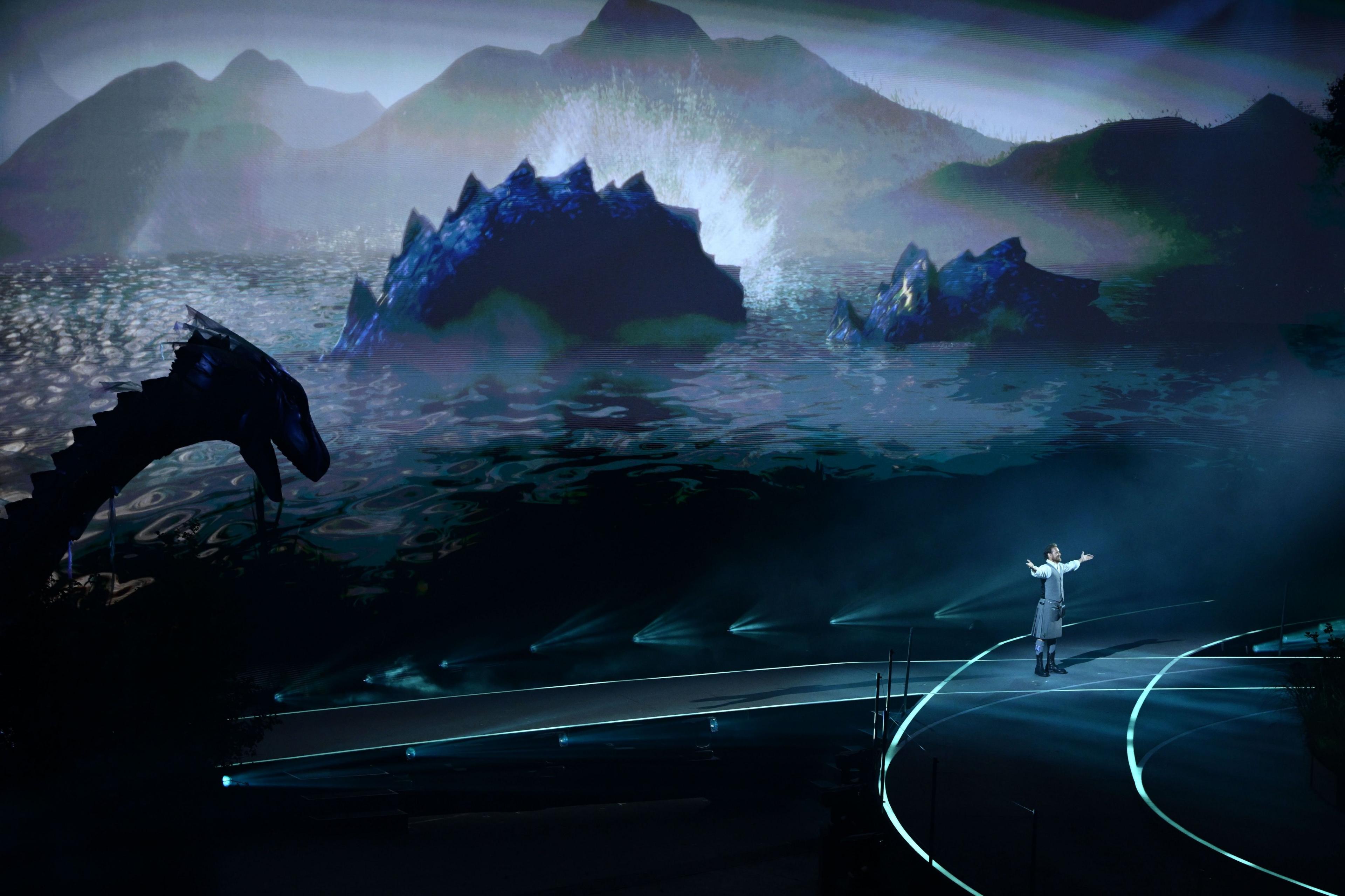 Scottish actor Sam Heughan performs next to Nessie. The stage is a dark blue, with Nessie's body projected onto a screen that shows water and mountains. Sam stands on the stage with his arms outstretched.