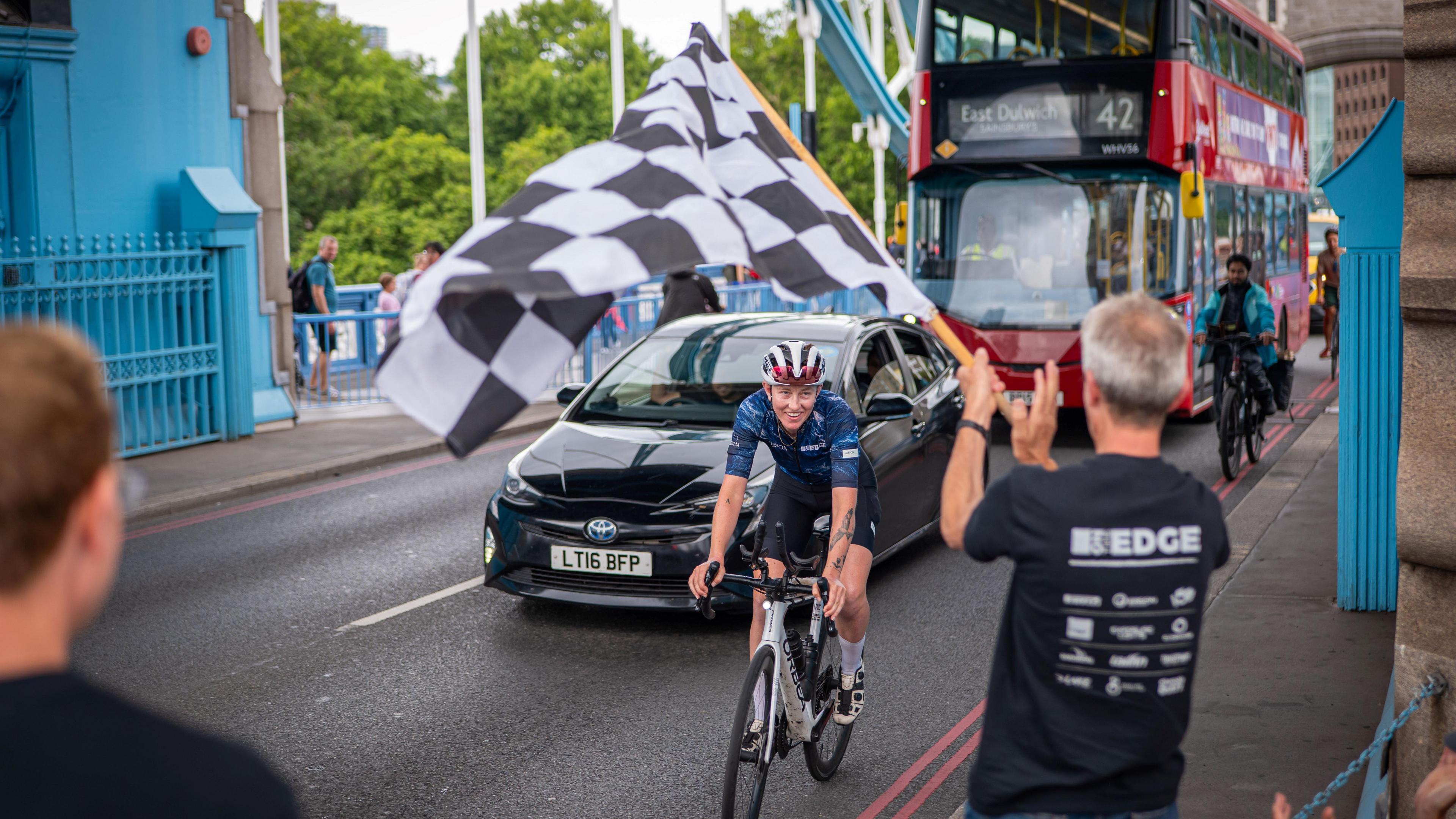 A woman rides on a bicycle towards us over a bridge, in front of a black car and red double decker bus. A man on the right is waving a black and white chequered flag.