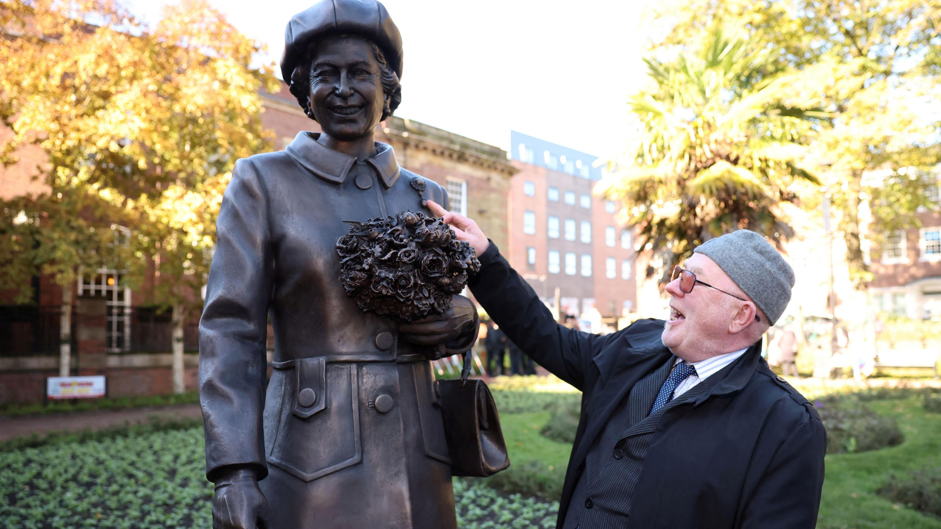 A man wearing a suit, glasses and a hat puts his right hand up to a bronze statue of Queen Elizabeth II.