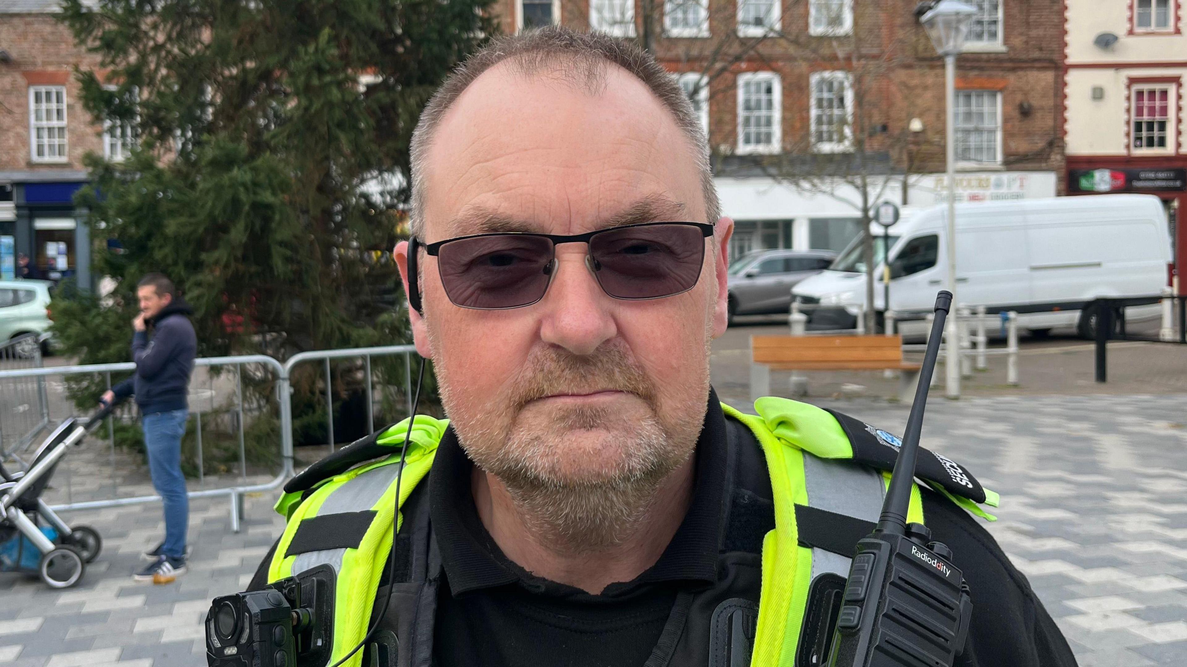 A man standing in a town square, with a Christmas tree behind him on grey paving slabs, and a row of shops behind that. The man has thinning brown hair and a greying bear and is wearing tinted glasses and a security guard uniform of a black top and hi-vis vest. He has a black walkie-talkie attached to his chest and a black earpiece in his right ear.
